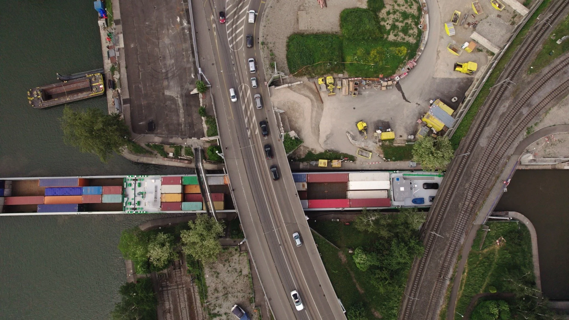 Luftaufnahme eines Flussufers, einer Brücke, eine Straße mit Autos, ein Frachtschiff, gebautes Gebiet mit Baustellen und Eisenbahnlinien.