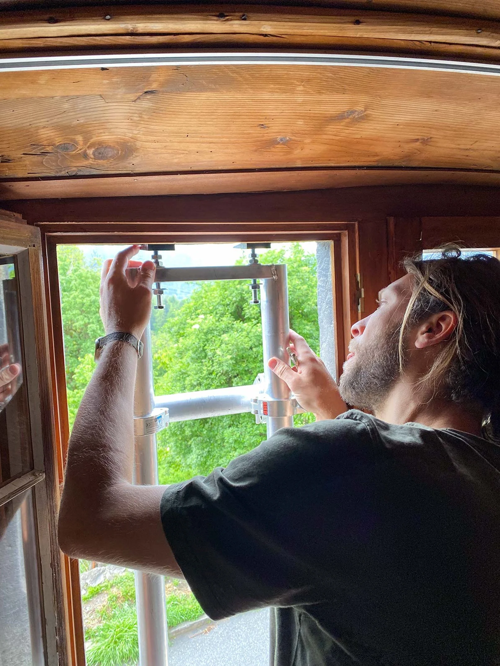 Ein Mann arbeitet an einem Fensterrahmen aus Holz und installiert einen Metallrahmen im Inneren, während es draußen grün ist.