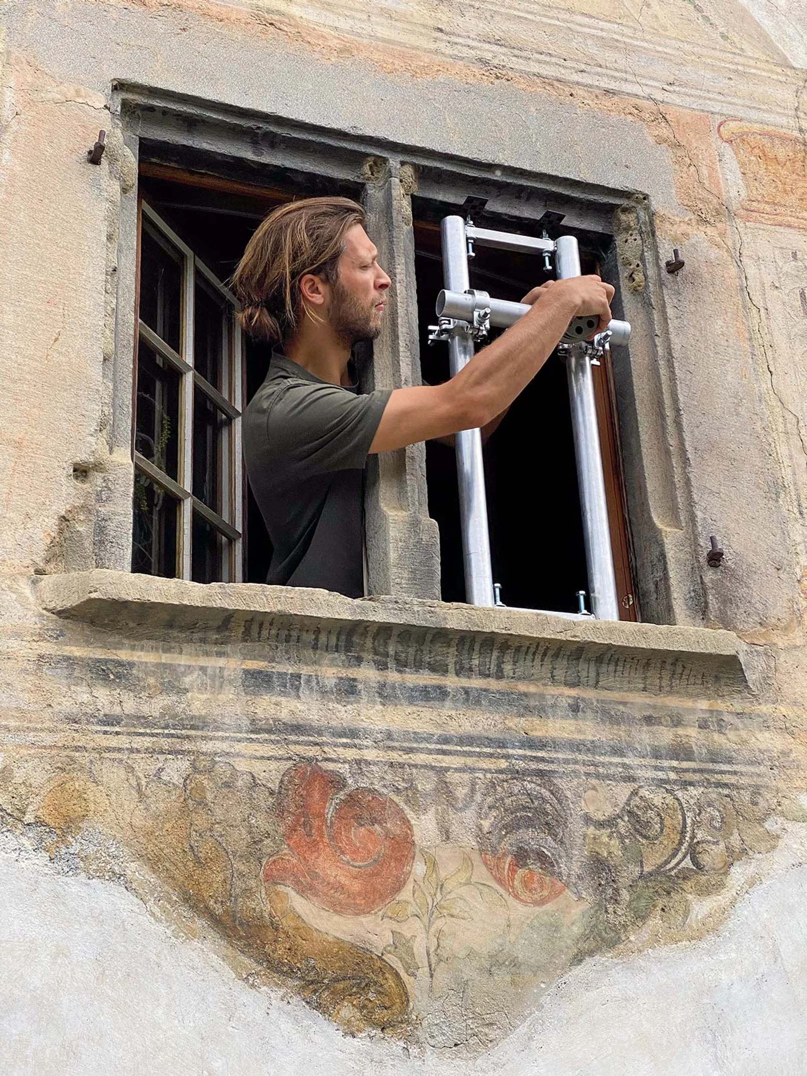 Ein Mann schaut durch ein Fenster mit Metallrahmen an einer alten, bemalten Wand.