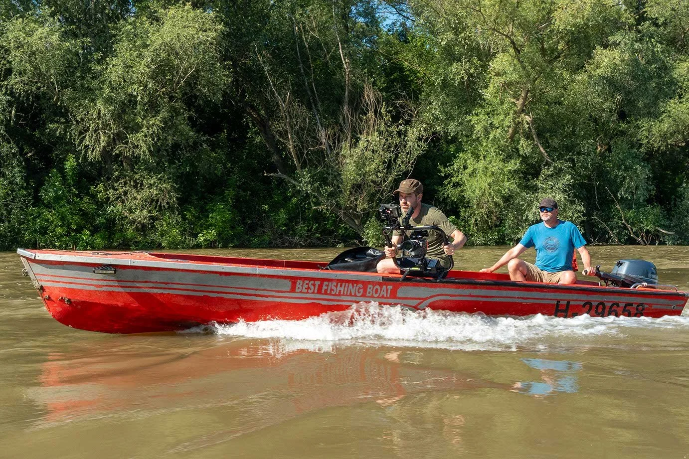 Zwei Männer sitzen in einem roten Boot auf einem Fluss, eines filmt mit einer Kamera. Das Boot ist mit 'Best Fishing Boat' beschriftet und hat die Kennzeichnung 'HF 29658'. Hinter ihnen sind grüne Bäume am Flussufer.