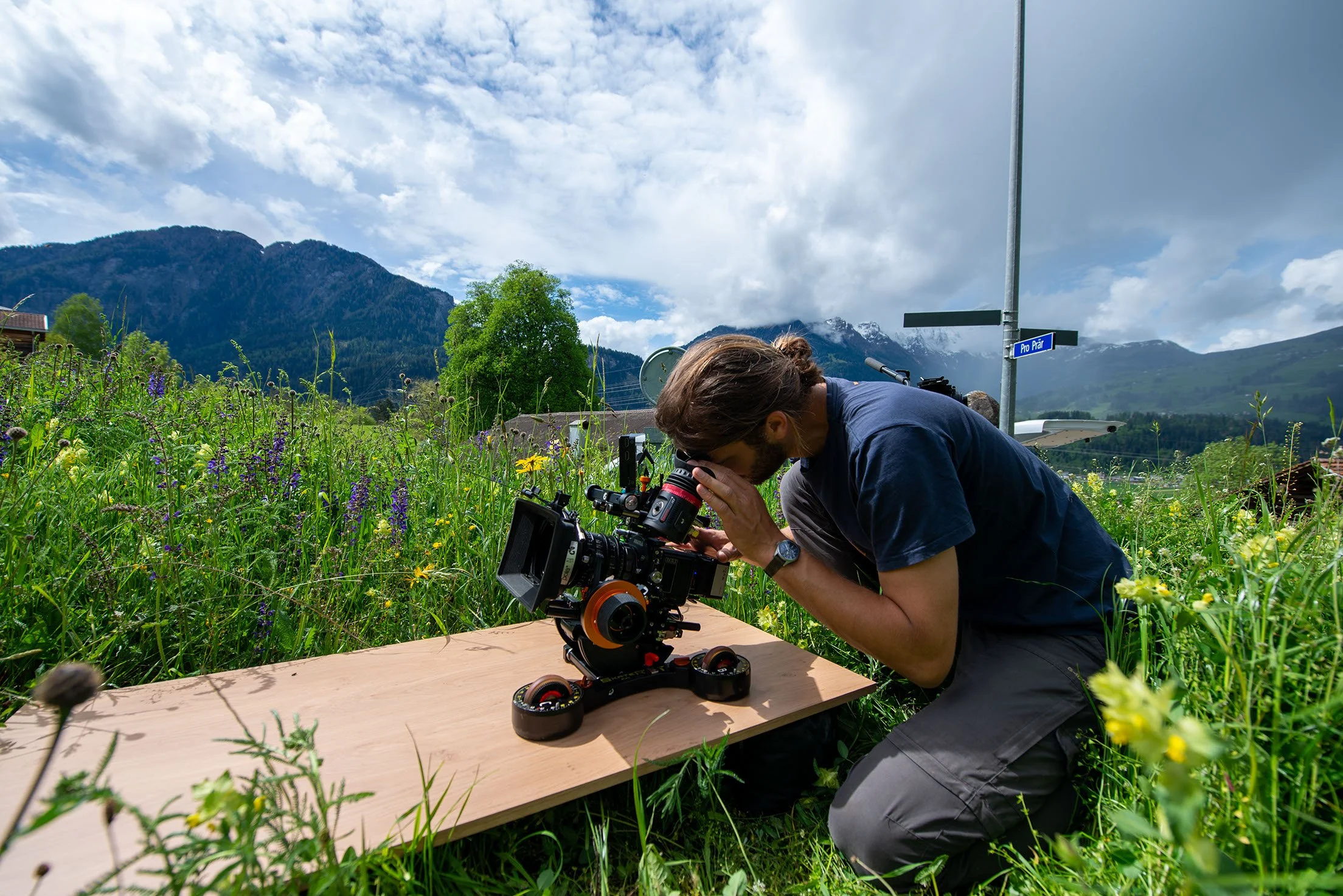 Ein Mann arbeitet mit einer Kamera auf einem Holzgestell im Freien, umgeben von Wildblumen und grünen Büschen, mit Bergen im Hintergrund und einem bewölkten Himmel.