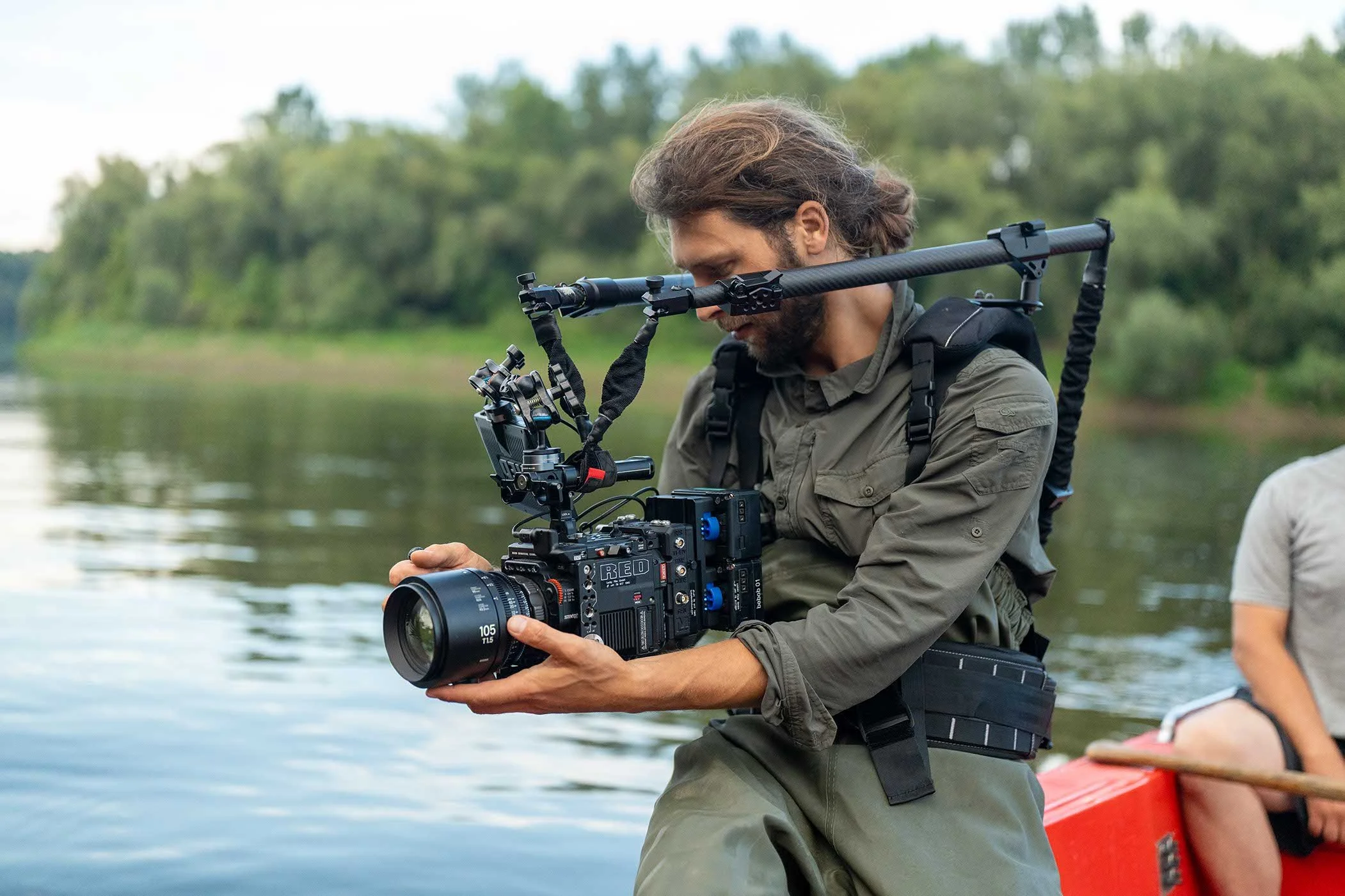 Ein Mann mit Kamera auf einem Boot am Fluss. Im Hintergrund Bäume und Wasser. Der Mann trägt eine grüne Jacke und einen Rucksack.