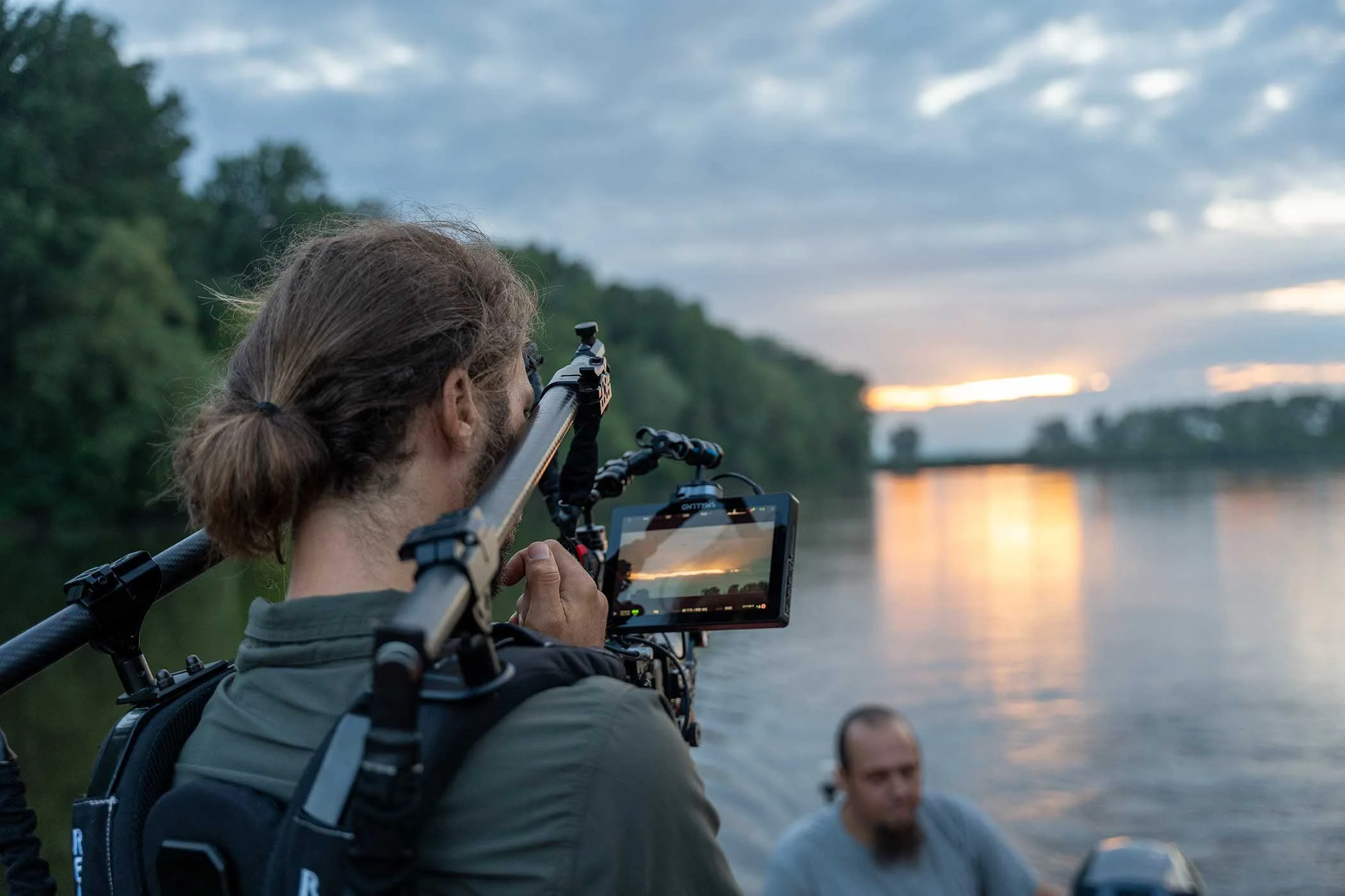 Ein Mann mit langen Haaren und Rucksack benutzt eine Kamera, um den Sonnenuntergang über einem Fluss zu filmen, während ein anderer Mann im Hintergrund zuschaut.