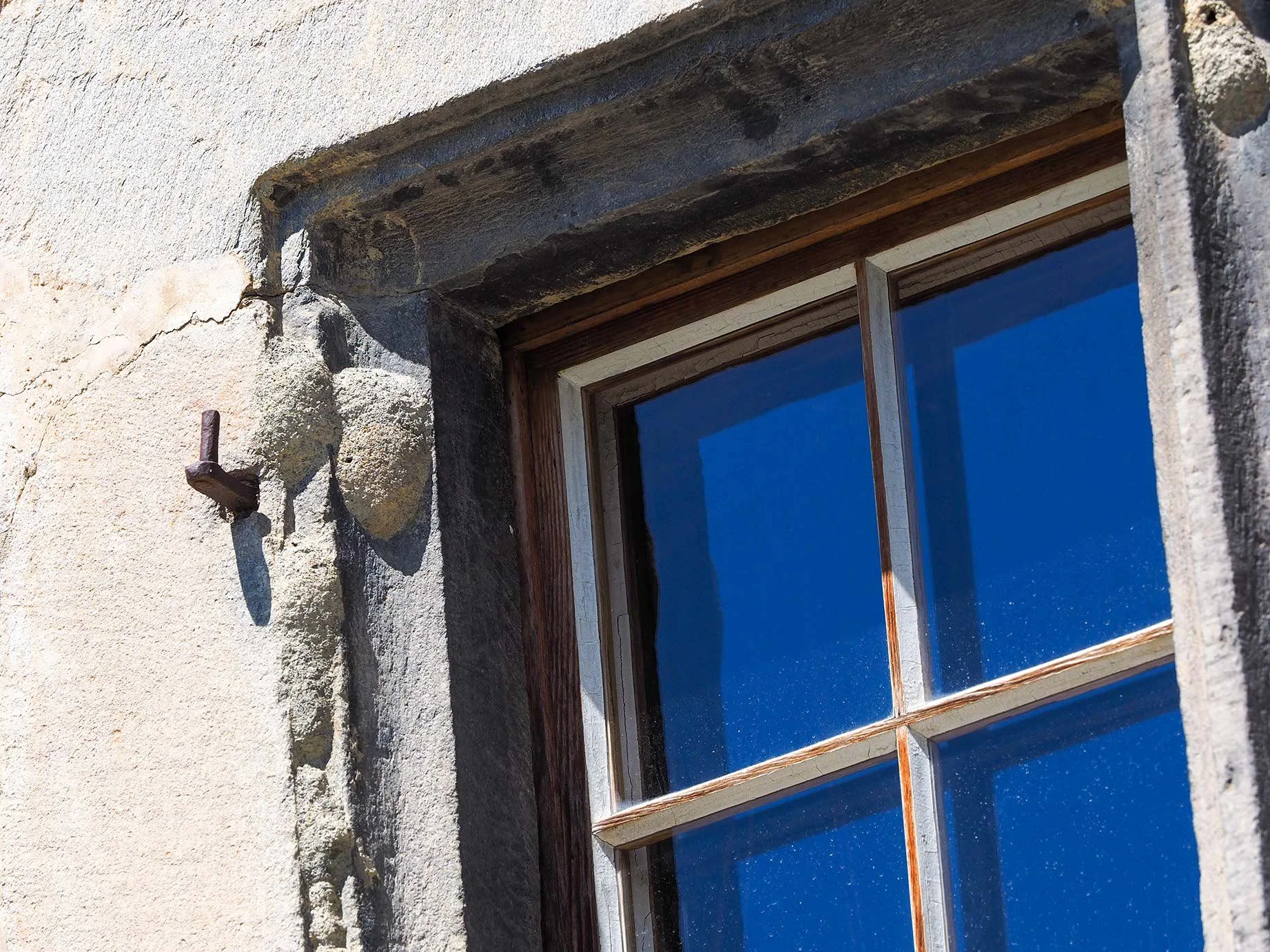 Nahaufnahme eines Fensters in einer alten Steinmauer, umgeben von grobem Putz. Das Fenster hat einen Holzrahmen und reflektiert den blauen Himmel.