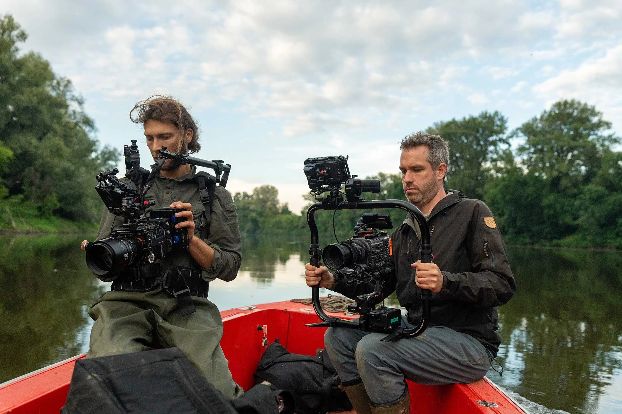 Zwei Männer, die in einem roten Boot auf einem Fluss sitzen, während sie professionelle Kameraausrüstung verwenden, um die Natur zu filmen, umgeben von Bäumen und einem bewölkten Himmel.