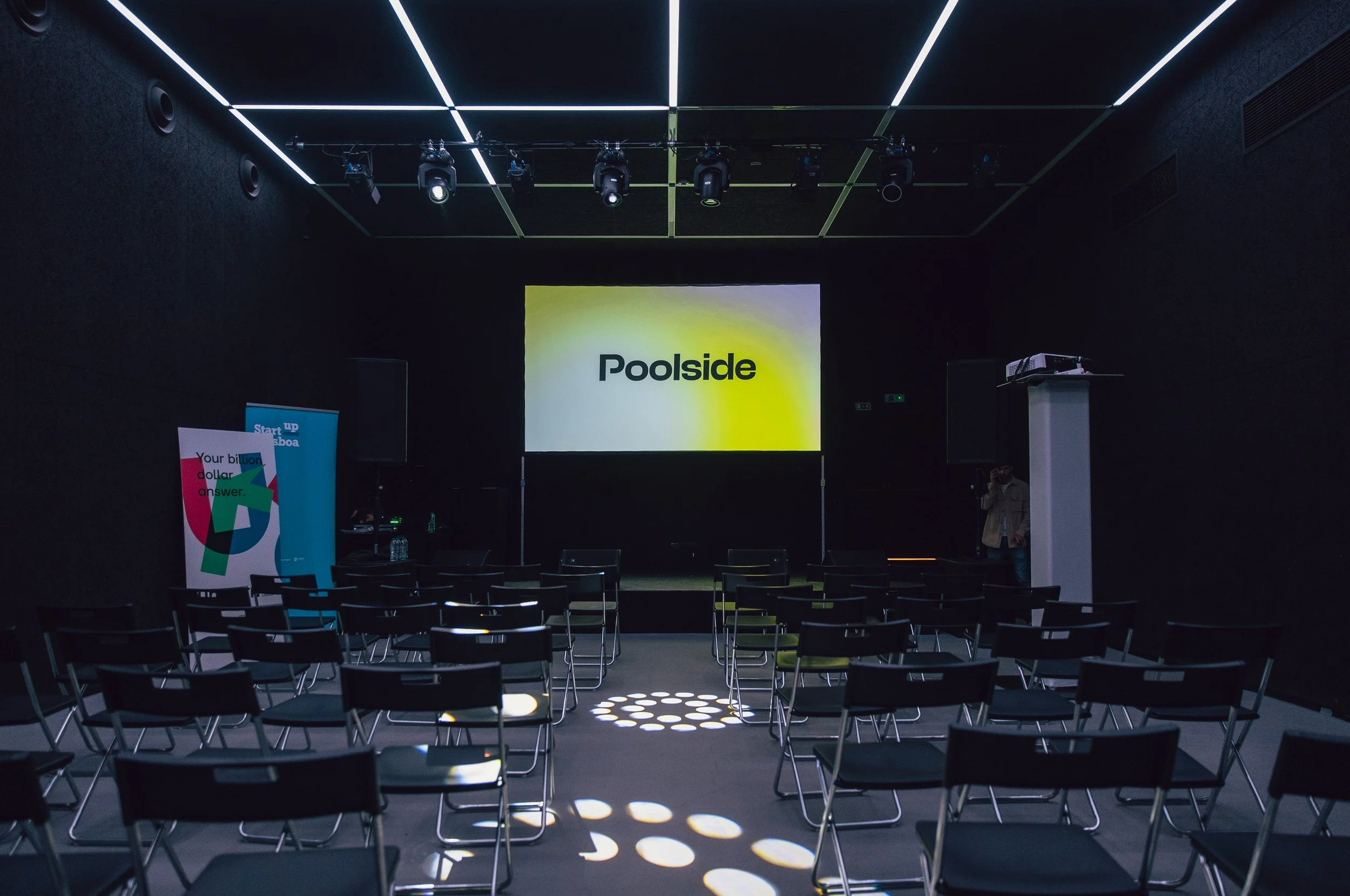 Sala multiusos privada do espaço Poolside, escura e insonorizada com cadeiras em fila para plateia, tela de projeção com o logo 'Poolside', banners laterais e iluminação moderna no teto.
