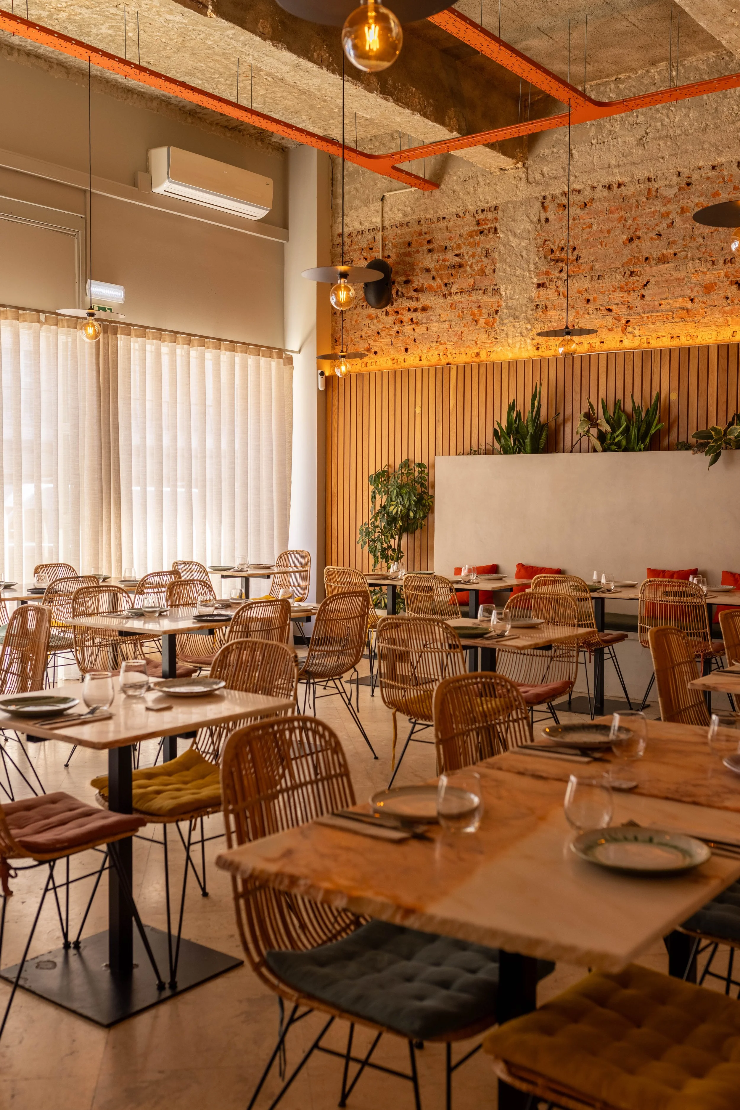 Sala principal do restaurante Cobaia em Alvalade, Lisboa com mesas de madeira, cadeiras de vime, cortinas claras, parede de tijolos expostos, iluminação aconchegante, plantas decorativas, criando ambiente luminoso e alegre.
