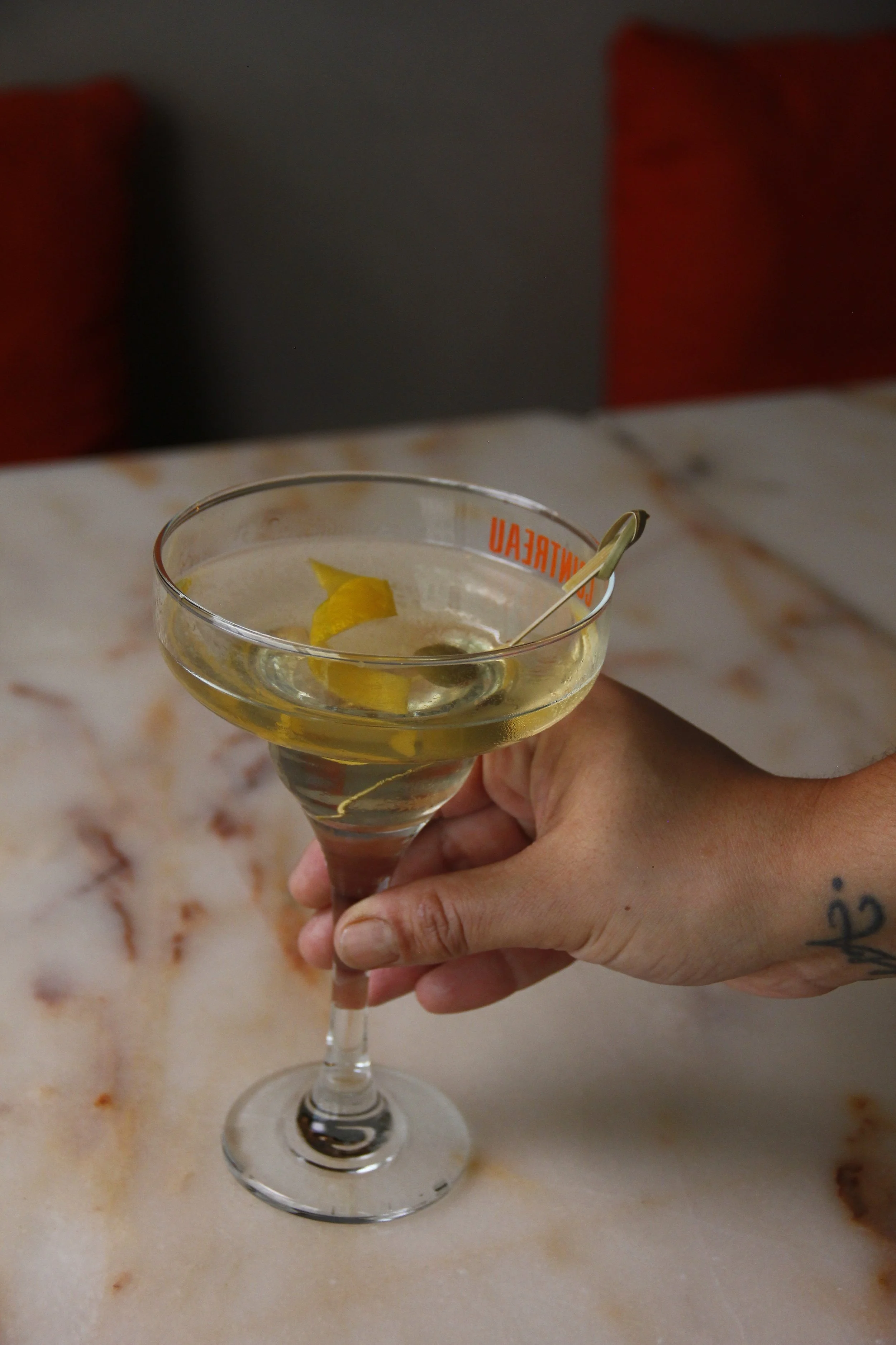 Pessoa segurando um cocktail de autor amarelo com limão do Restaurante Cobaia em Alvalade, Lisboa, num copo alto, sobre mesa de mármore.