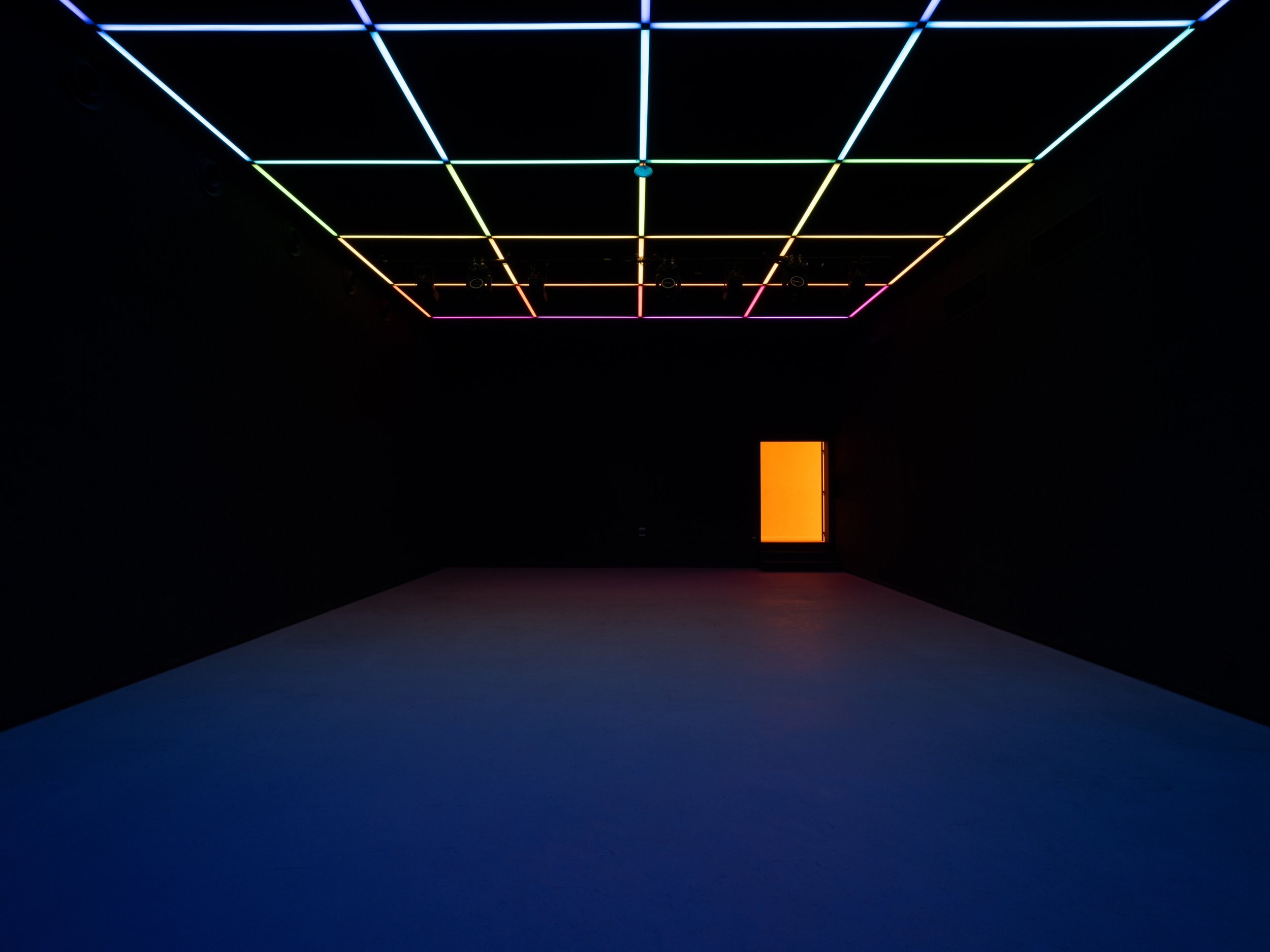 Sala multiusos do espaço Poolside, escura com teto de linhas de luz neon coloridas formando uma grade, uma entrada laranja iluminada ao fundo da sala e piso escuro.