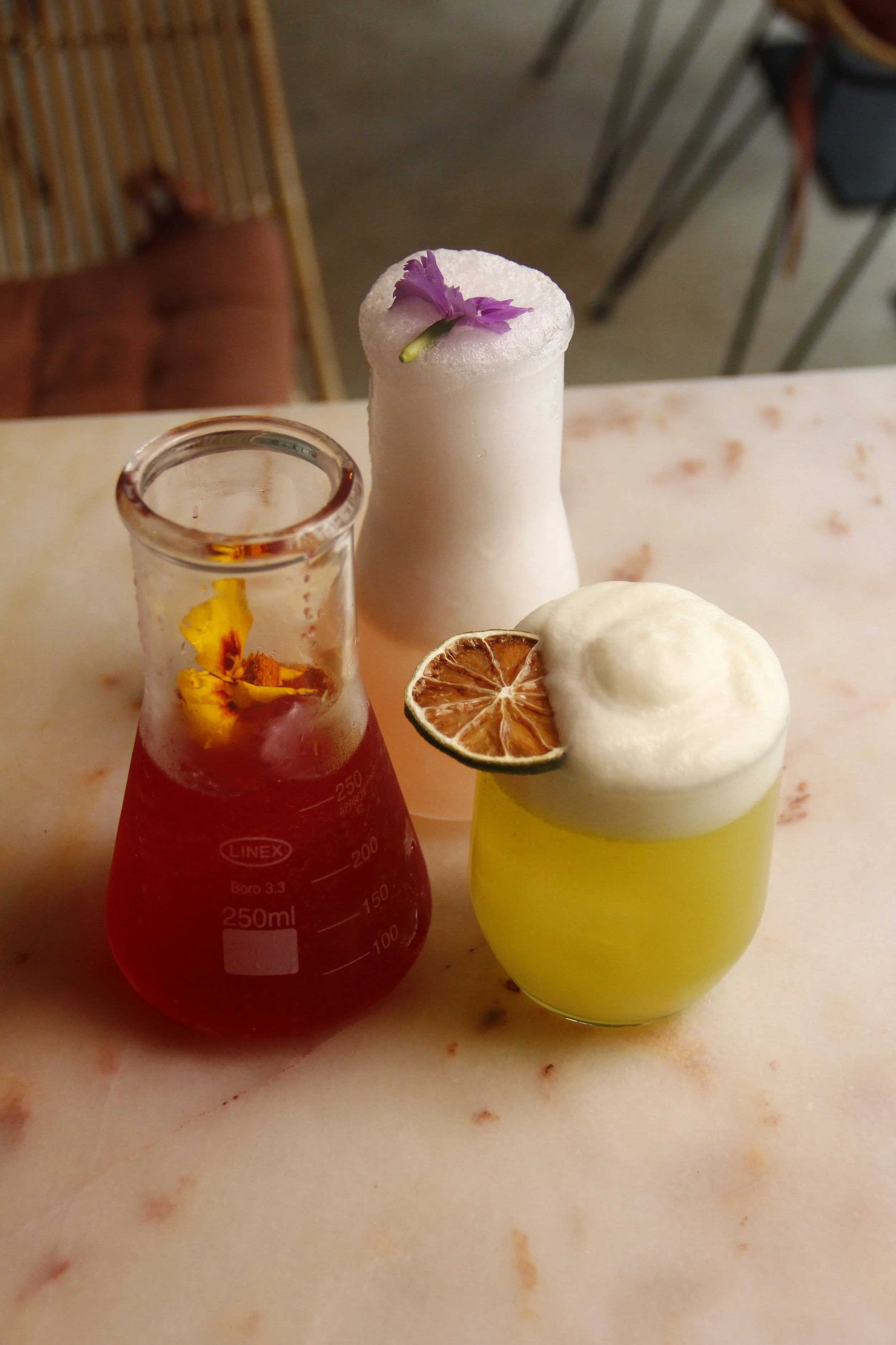 Três cocktails de autor do Restaurante Cobaia em Alvalade, Lisboa coloridos e frescos com flores decorativas sobre uma superfície de mármore no ambiente de um café ou bar.