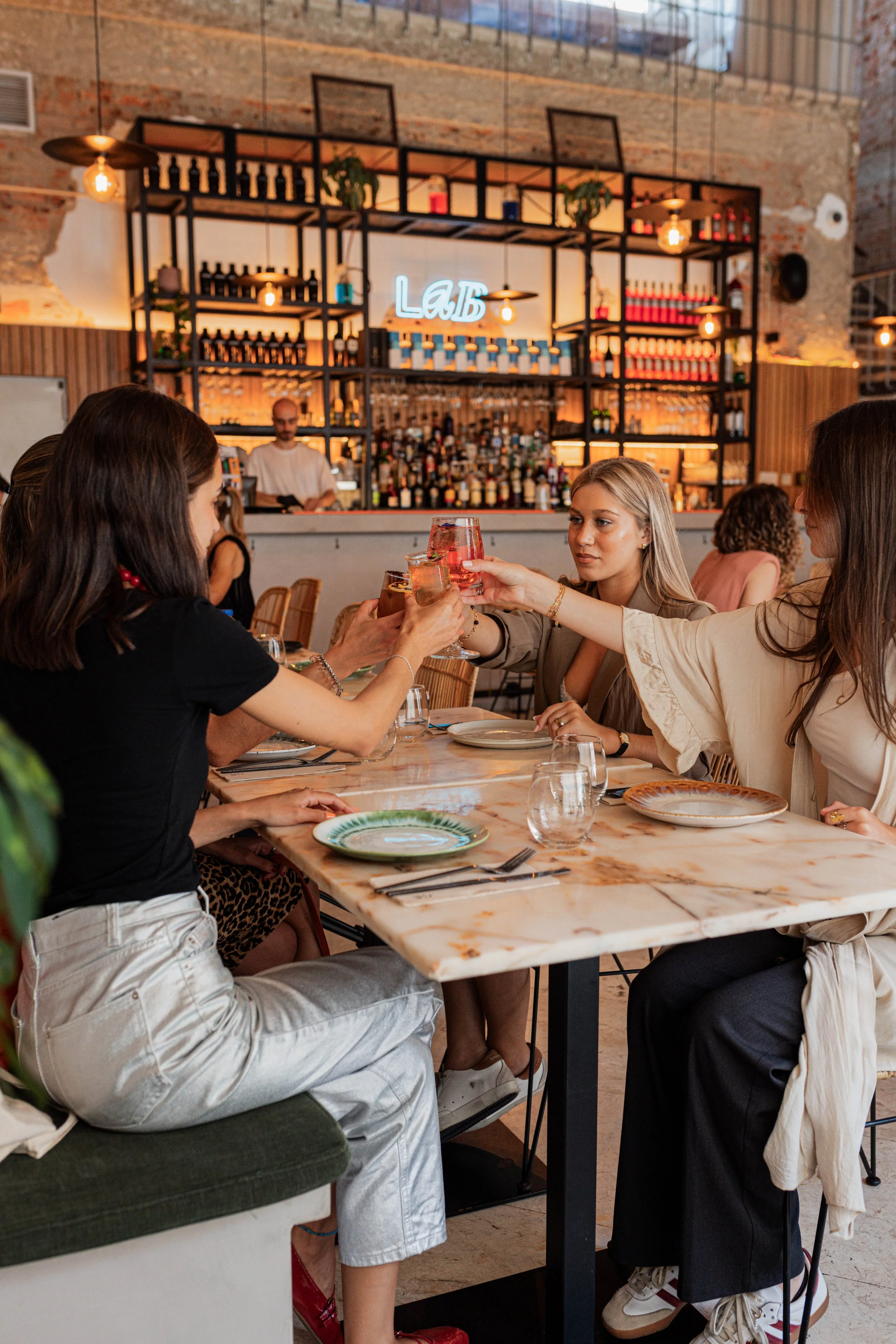 Almoço de grupo à mesa na sala principal do Restaurante Cobaia em Alvalade, Lisboa a brindar com bebidas com o bar e bebidas ao fundo.