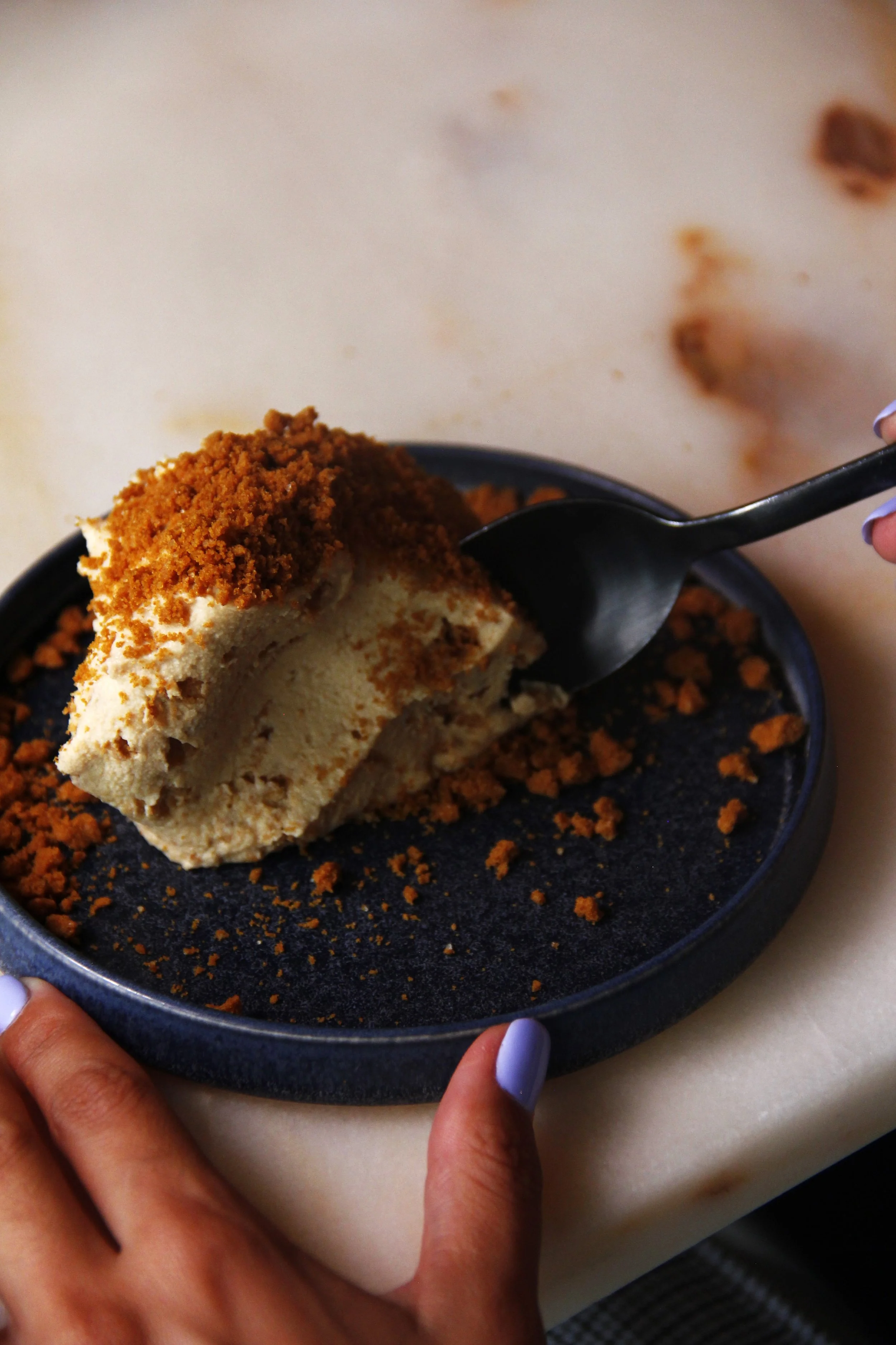 Close-up de pessoa a provar sobremesa Mousse Cobaia, com bolacha Lotus, do Restaurante Cobaia em Alvalade, com aspeto cremoso e cobertura de bolacha, servida num prato preto sobre mesa clara.