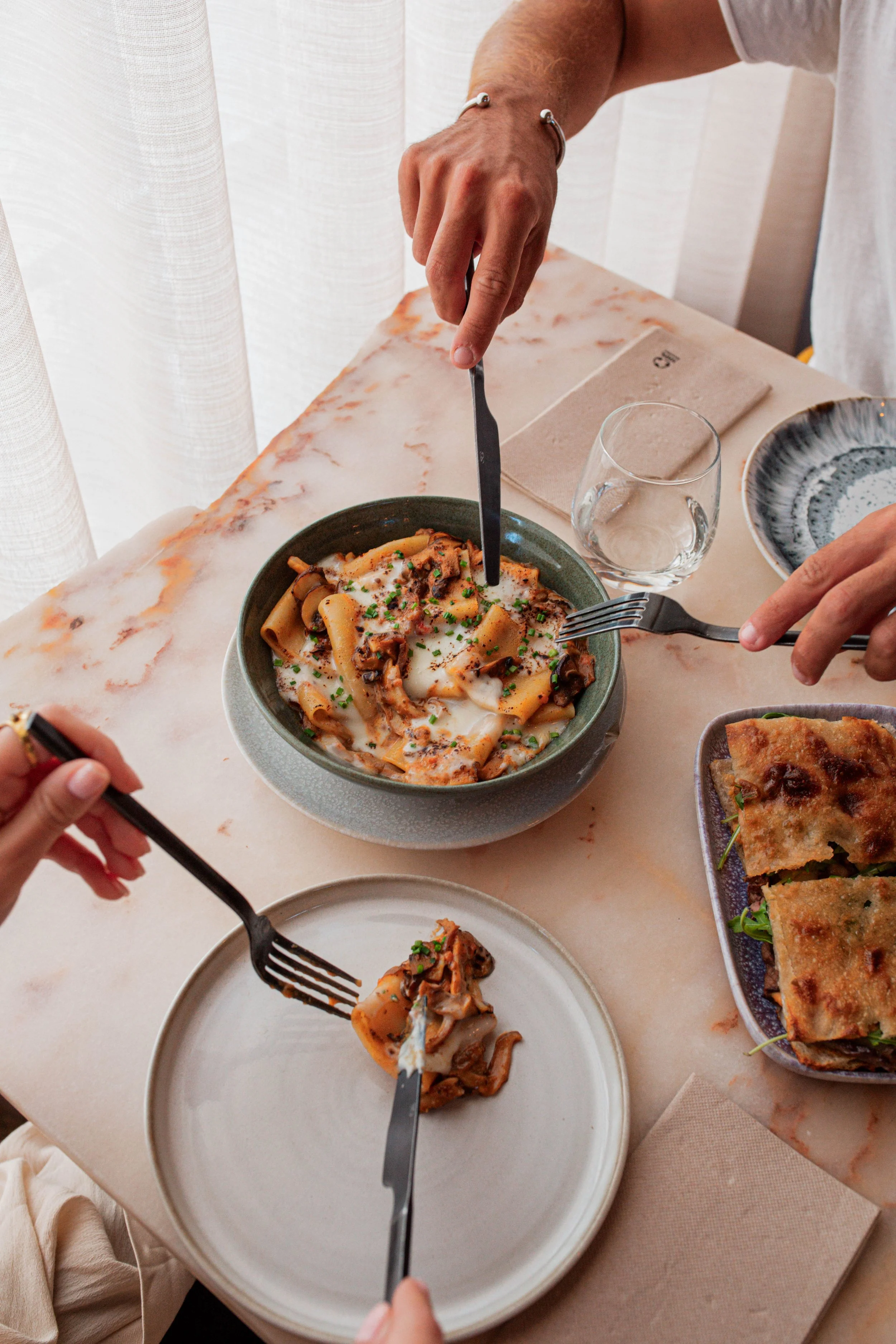 Pessoas compartilhando uma refeição de ragu de cogumelos vegetariano do restaurante Cobaia em Alvalade, Lisboa com aspeto delicioso numa mesa de mármore claro.