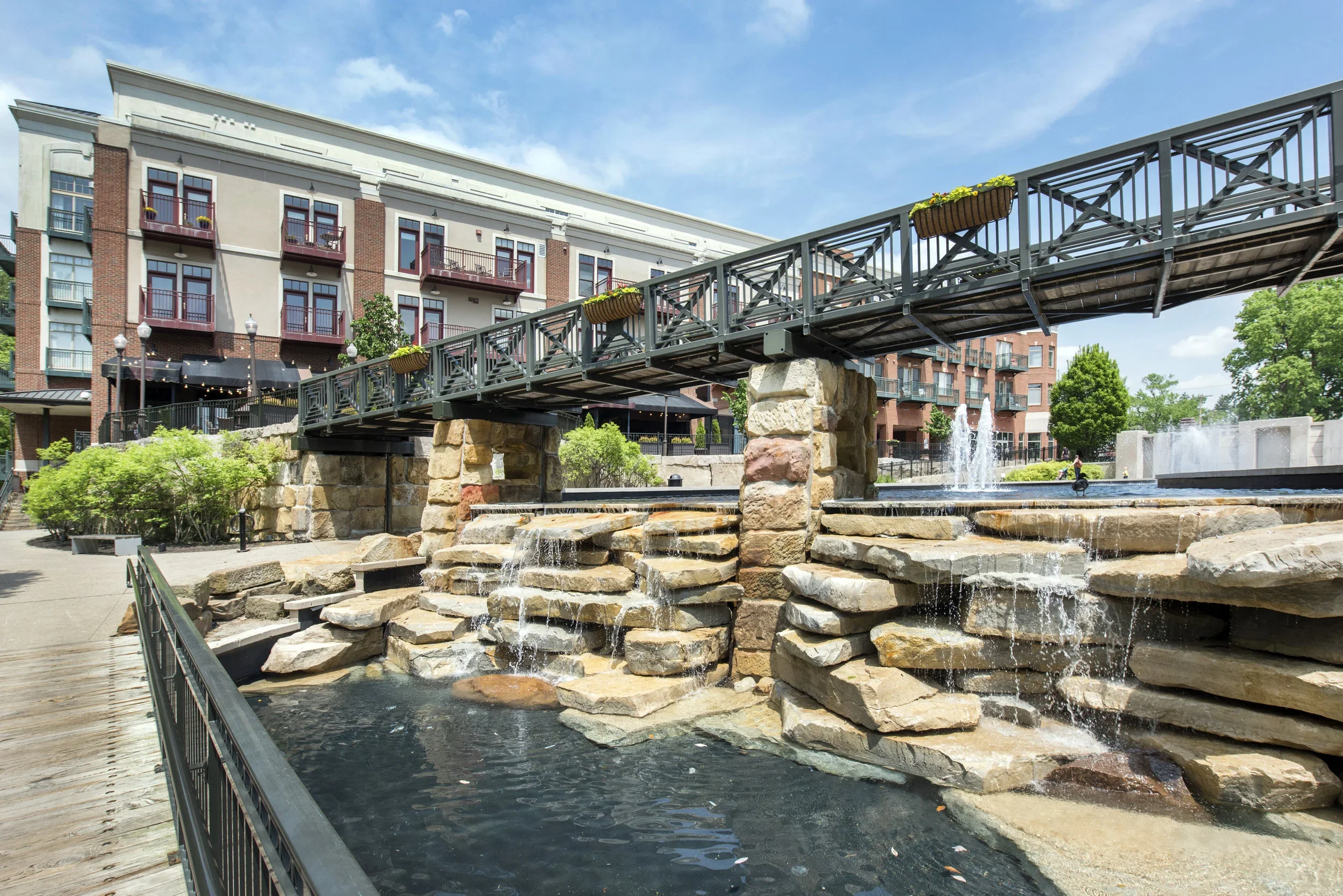 A city park with a waterfall feature, rocks, a bridge with planters, and apartment buildings in the background.