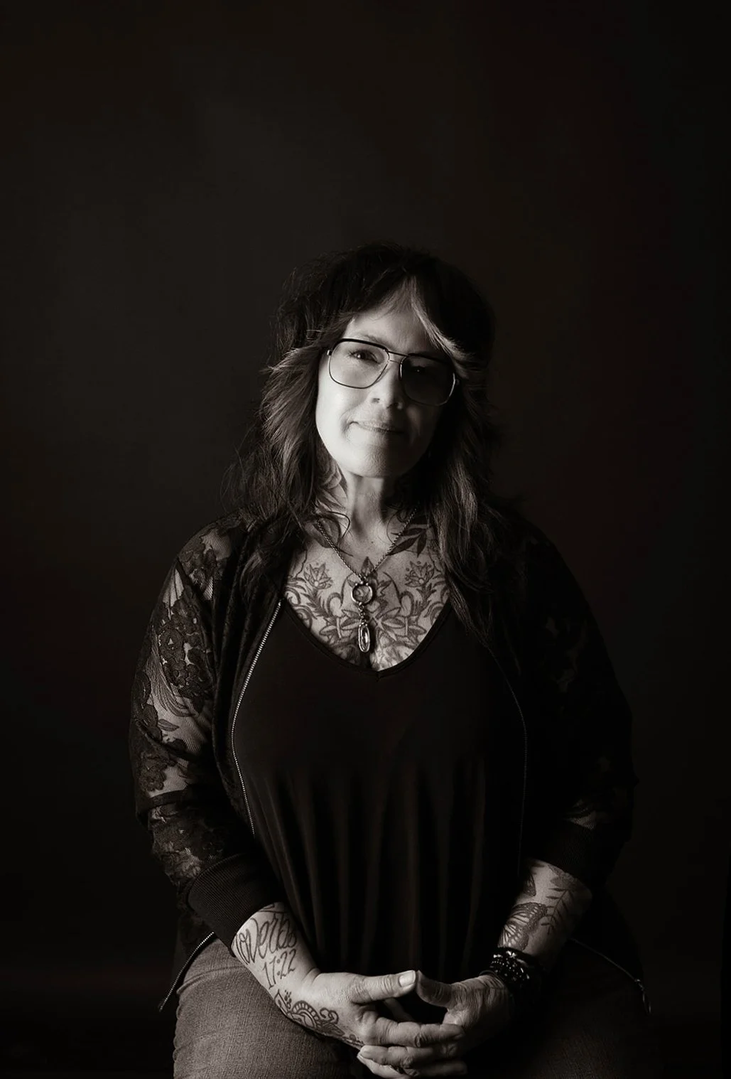 A woman with long wavy hair, glasses, and tattoos on her arms and chest, wearing a black top with lace sleeves and a necklace, sitting against a dark background.