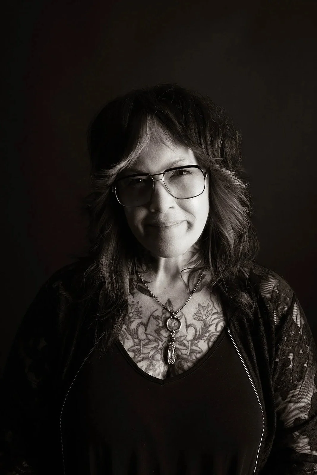 A black and white photo of a woman with wavy hair, glasses, and tattoos on her chest, wearing a necklace and a lace jacket.