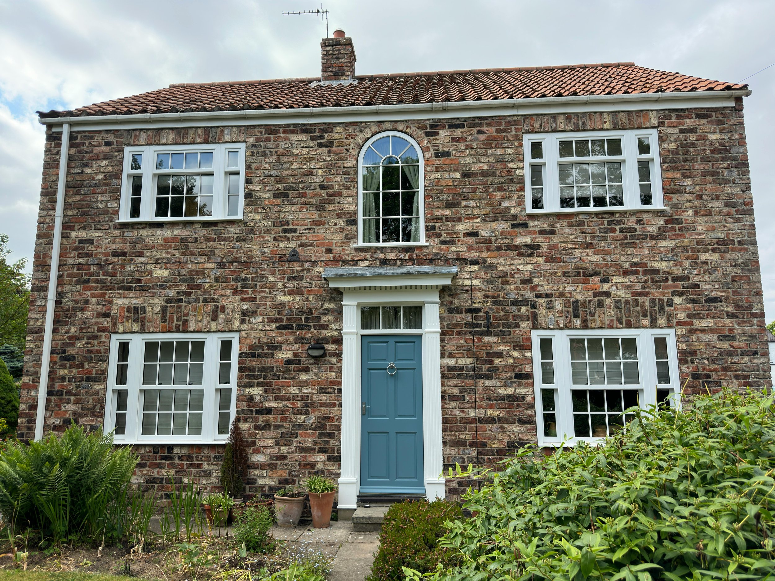 Brick house with white-framed windows and a blue front door, surrounded by greenery.