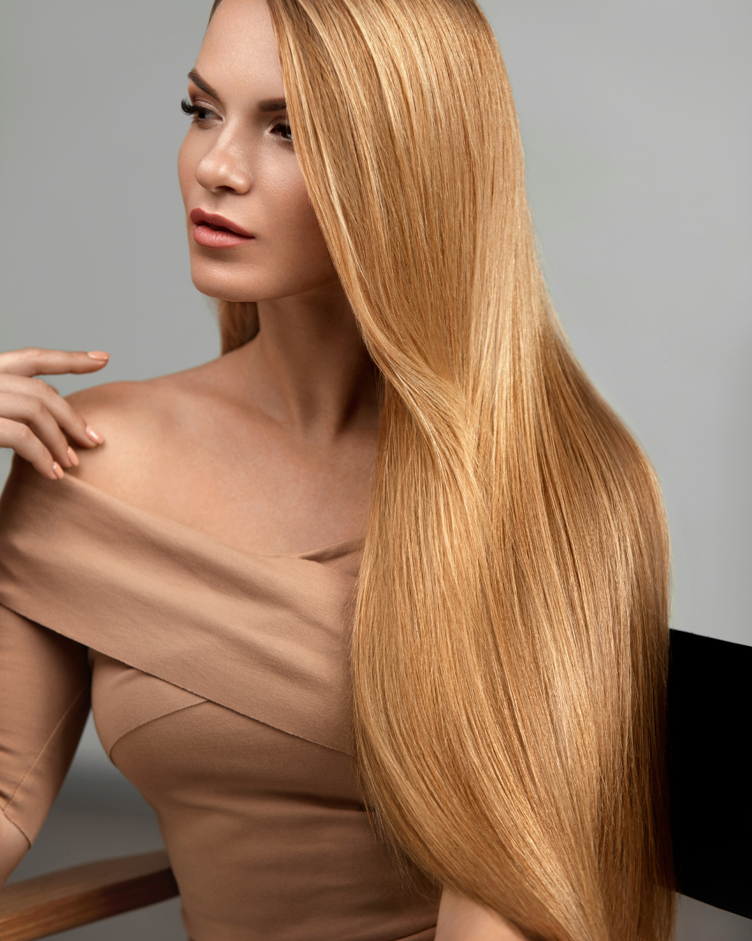 A woman with long, straight, blonde hair looking to the side, wearing a beige off-the-shoulder top, sitting against a neutral background.