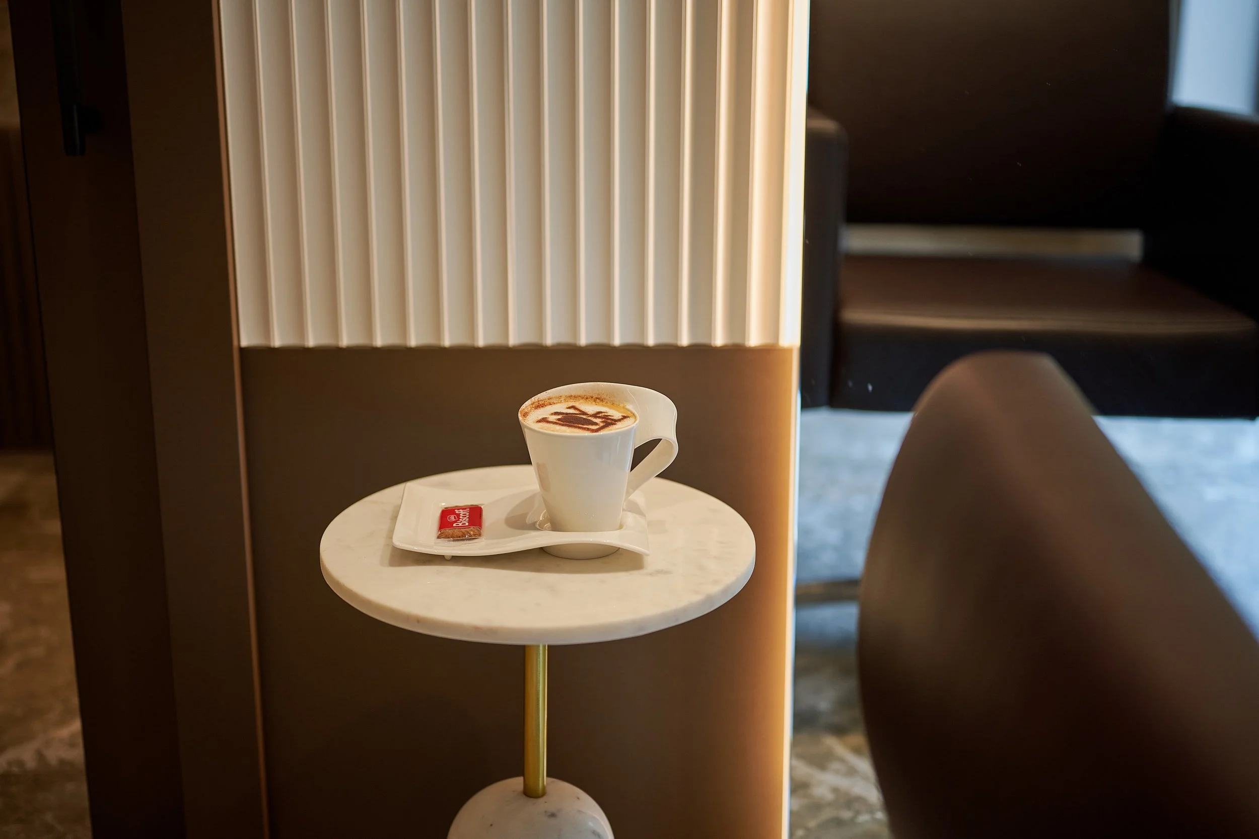 A white coffee cup with a hot beverage, possibly cappuccino or latte, on a small marble round table. The cup has a cappuccino art design on top. There is a small packet of sugar on a white saucer beneath the cup. The background includes modern decor