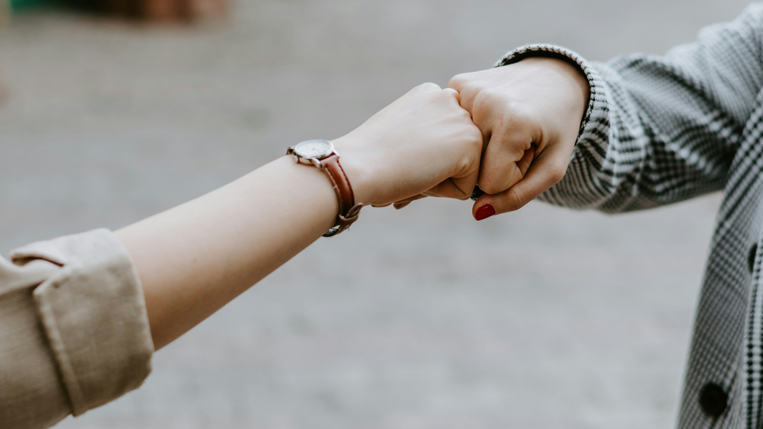 Two people are holding hands, with one person's hand resting on top of the other's in an outdoor setting. One person is wearing a checkered long sleeve shirt and a watch with a brown leather strap. The background is blurred.