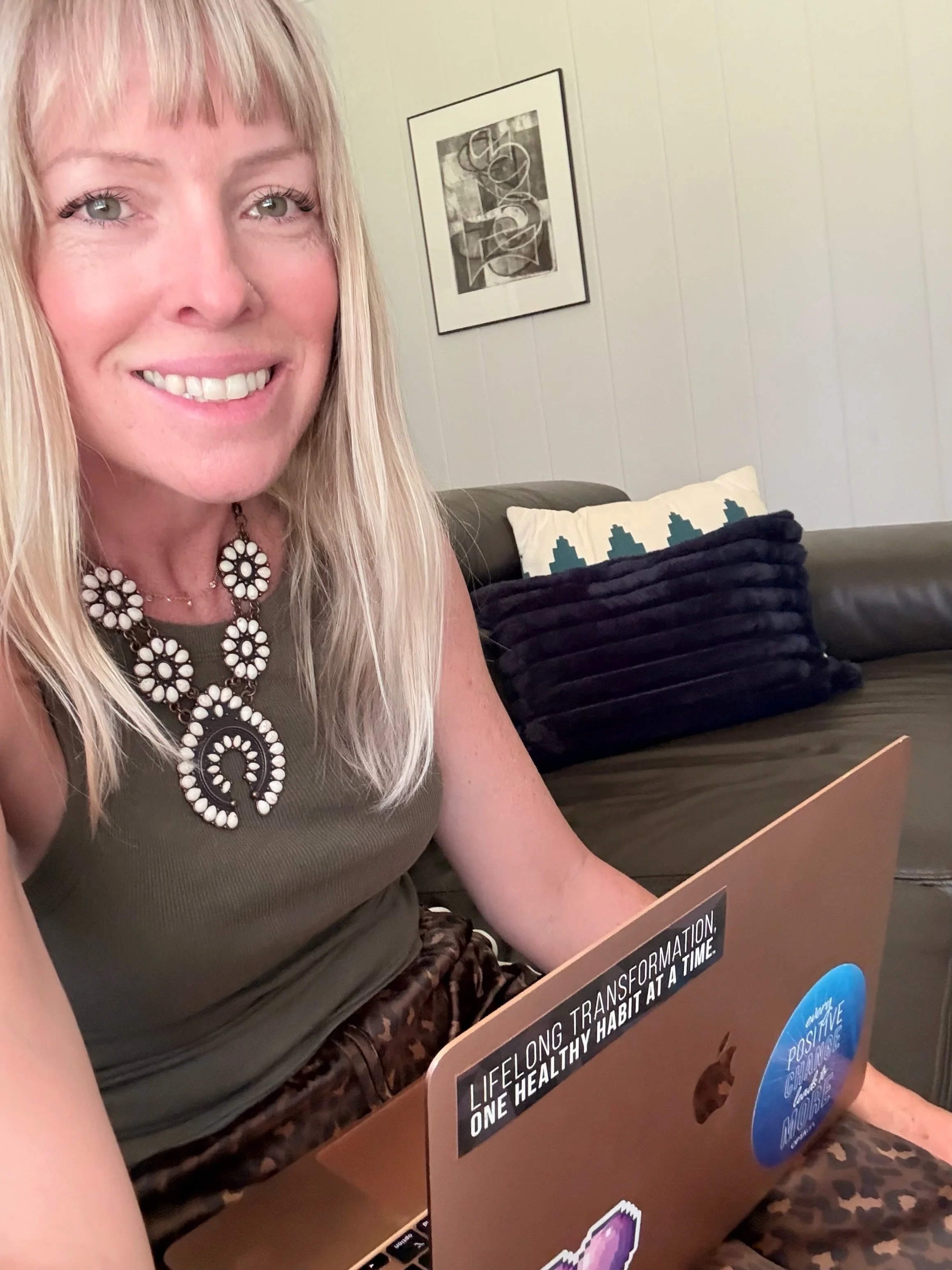 A woman with blonde hair wearing a statement necklace sitting on a couch, working on a laptop, with a pillow and some artwork on the wall behind her.