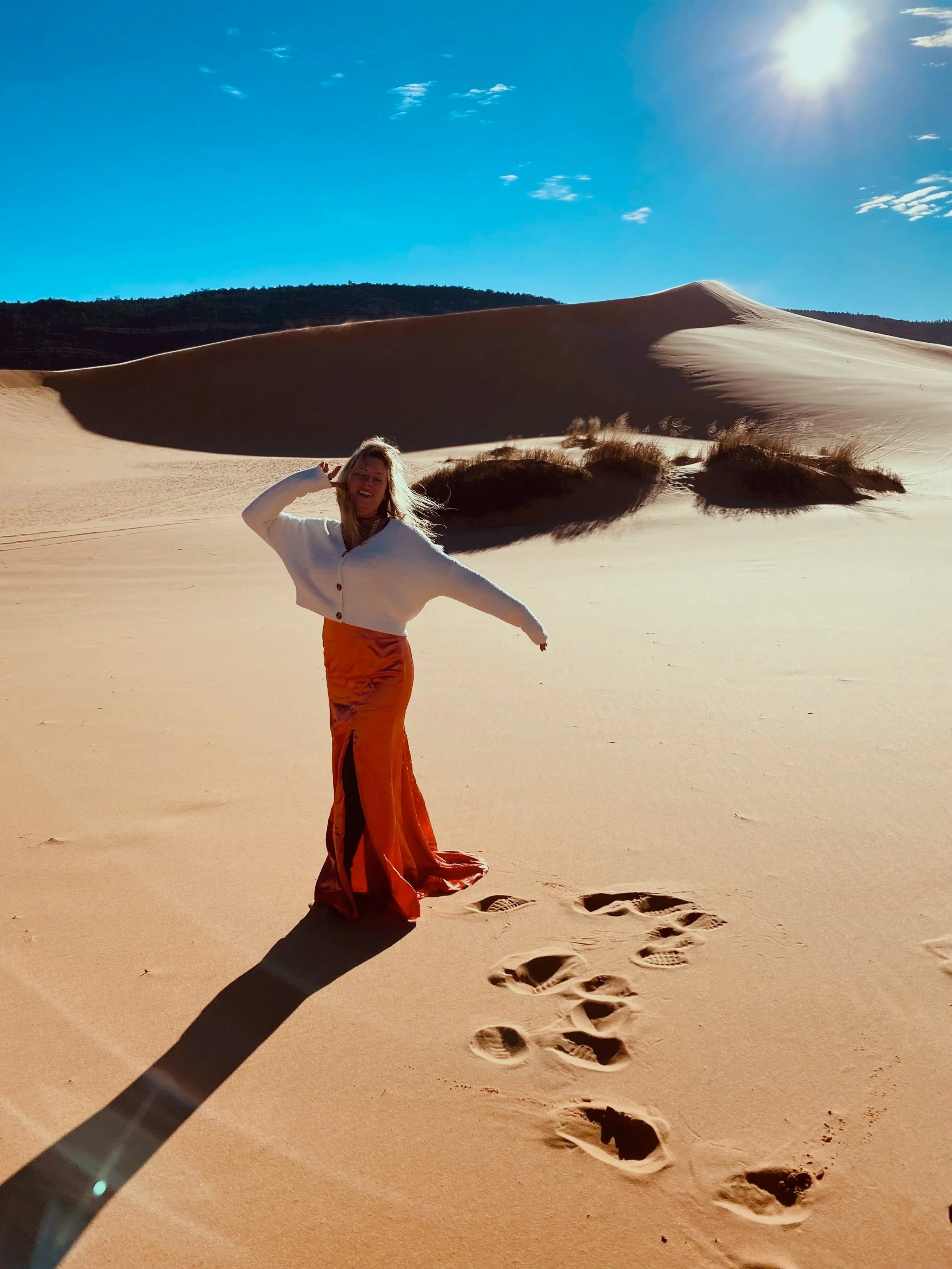 A woman standing in a desert with large sand dunes, wearing a white cardigan and an orange skirt, smiling and making a playful gesture. Footprints are visible in the sand near her. The sky is clear with a few clouds and the sun shining brightly.