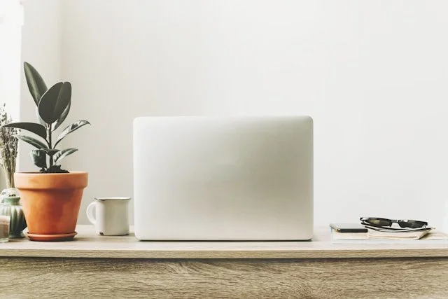 Laptop on a wooden desk with a plant in a terracotta pot, a white mug, and a pair of glasses on a pad of paper.