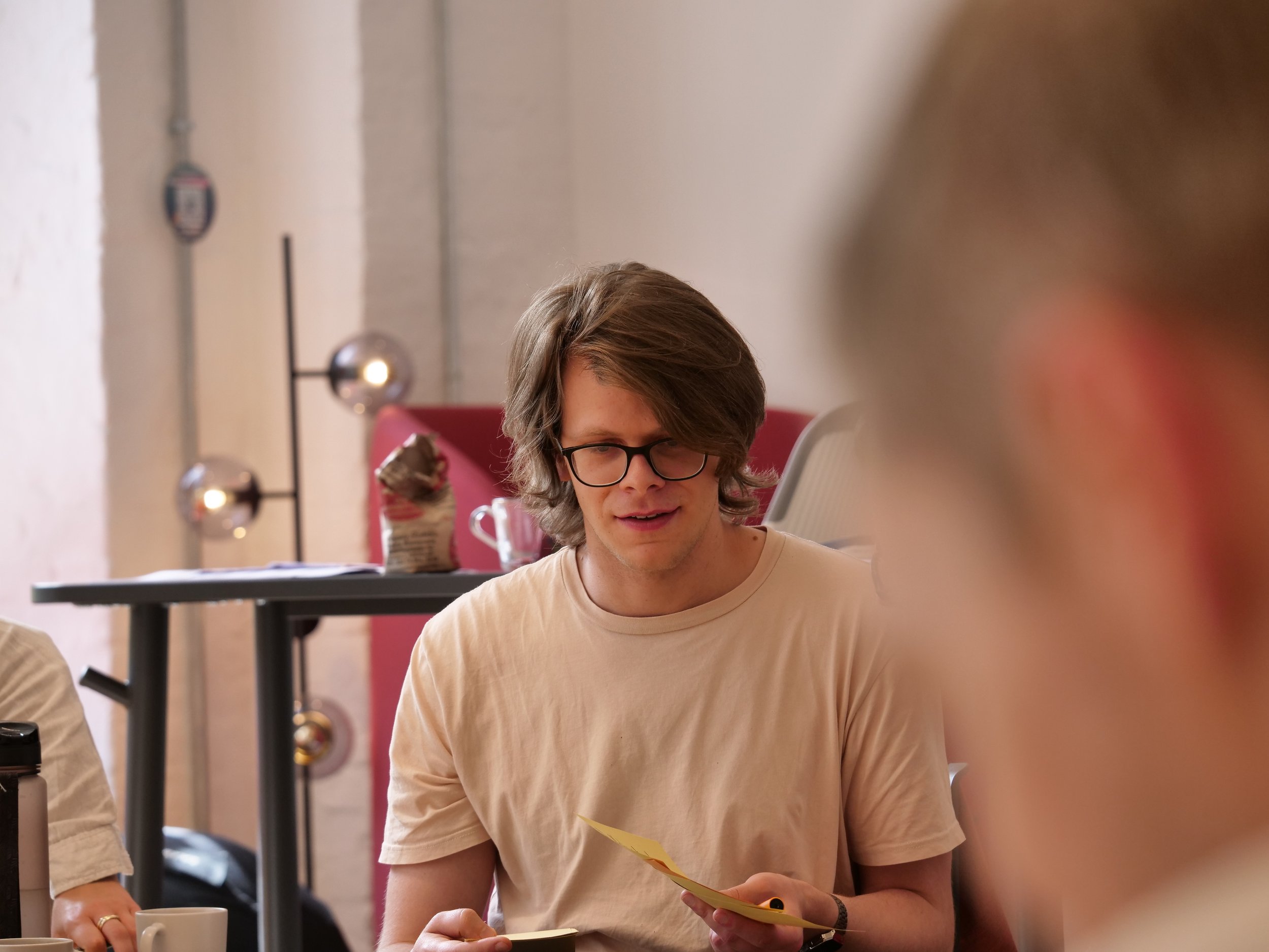 Ein junger Mann mit Brille und mittellangem braunem Haar liest einen Zettel, sitzt an einem Tisch in einem Raum mit modernen Lampen und Pastelltönen.