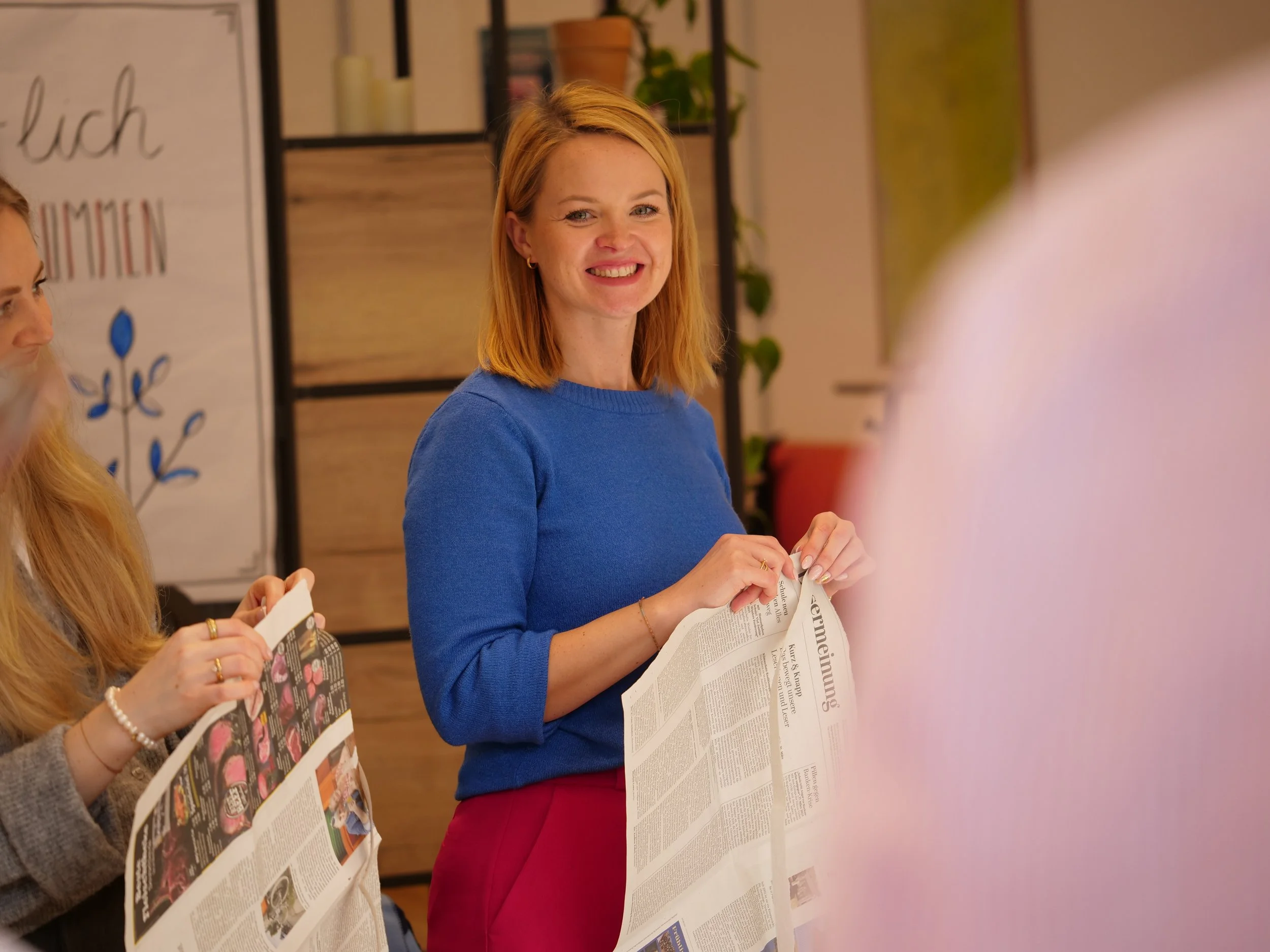 Eine lächelnde Frau mit roten Haaren trägt eine blaue Bluse und hält eine Zeitung. Sie ist in einem Raum mit Pflanzen und einer Tafel im Hintergrund.