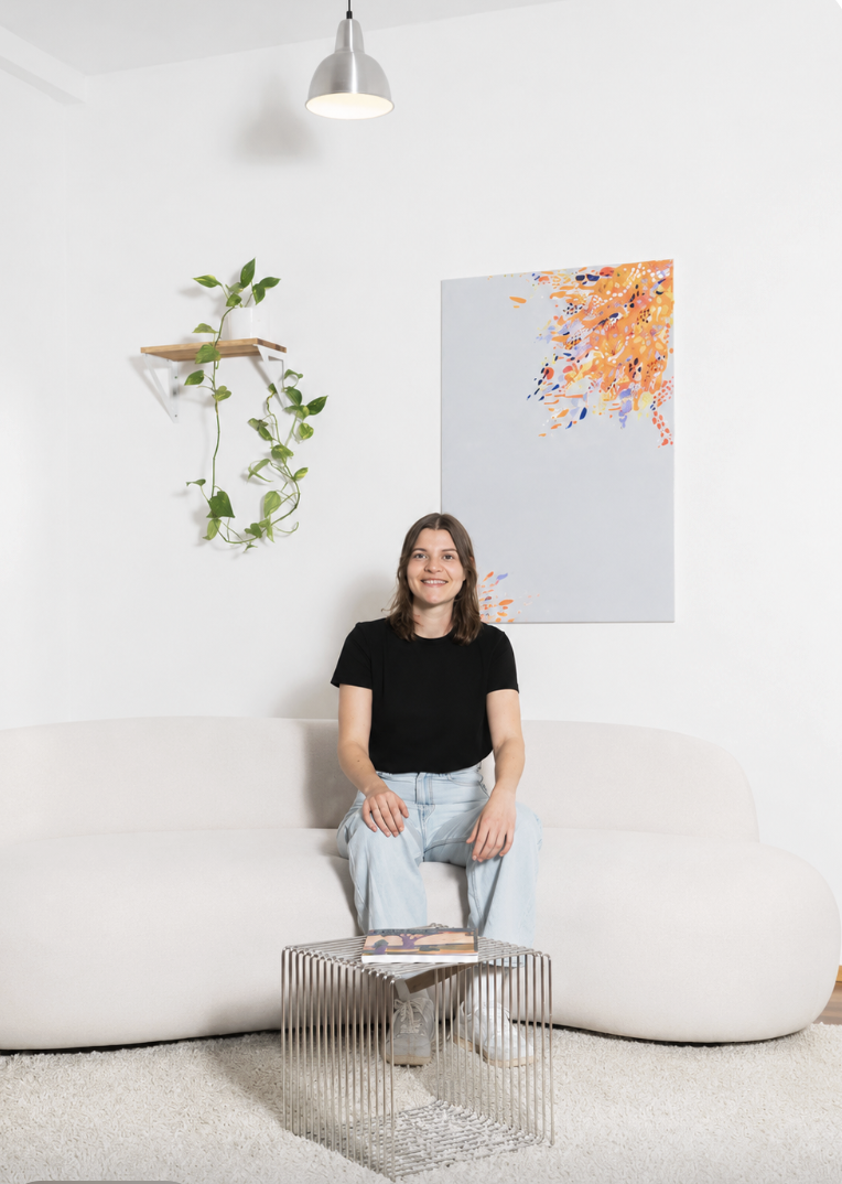 Junge Frau sitzt auf einer weißen Couch in einem modernen, hellen Raum, hinter ihr ein abstraktes Gemälde mit orange, blaue und lila Farben, an der Wand hängt eine Pflanze auf einem kleinen Holzregal, eine Deckenlampe hängt über ihr, ein Metallbeistelltisch steht vor ihr mit einem Magazin darauf.