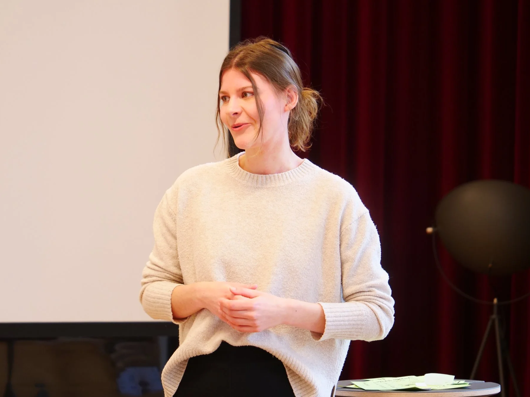 Eine junge Frau mit braunem Haar trägt einen cremefarbenen Pullover und steht in einem Raum mit einem weißen Bildschirm im Hintergrund und einem schwarzen Lautsprecher. Sie scheint zu sprechen.