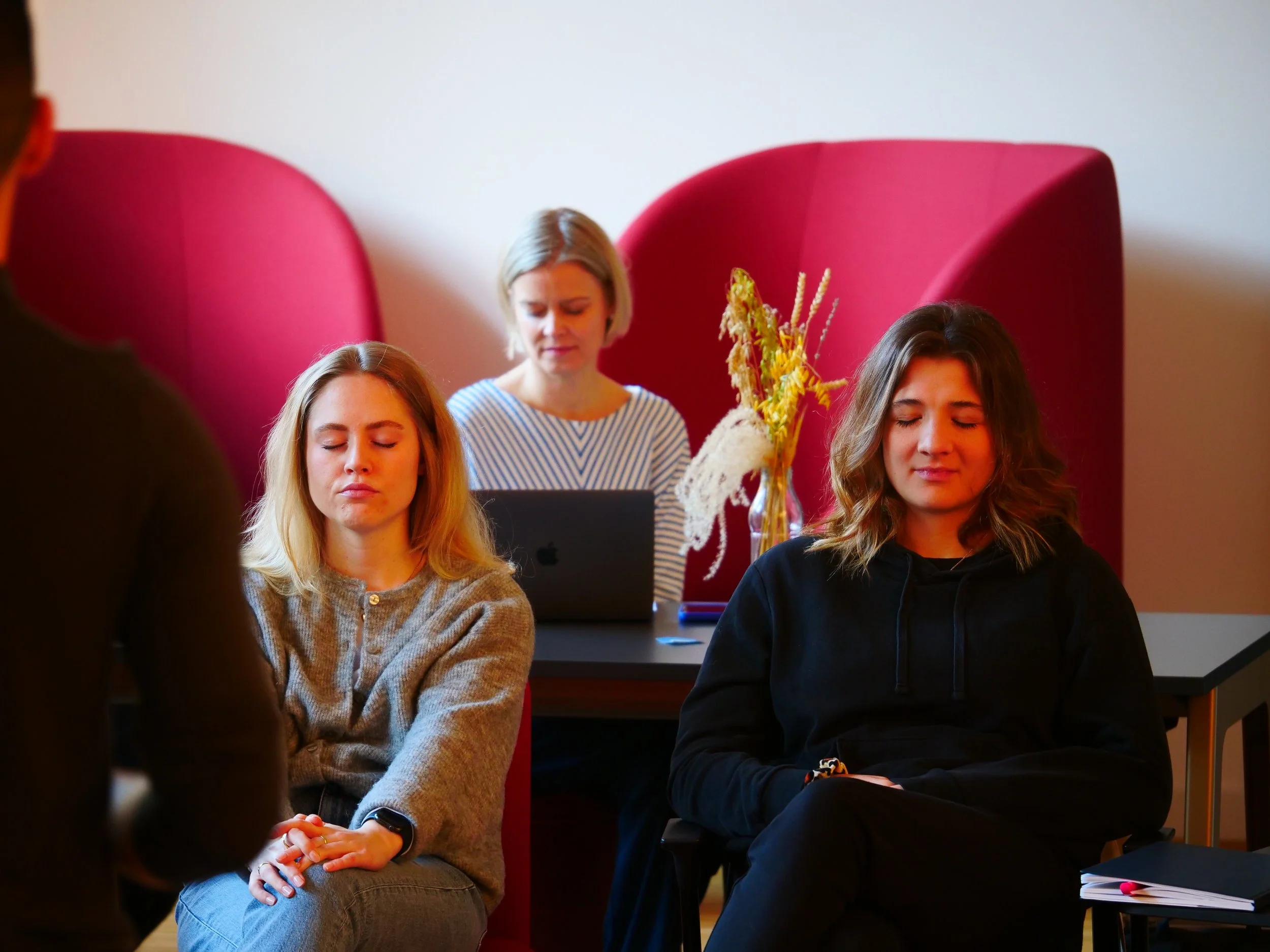 Drei junge Frauen sitzen im Meditationskurs mit geschlossenen Augen, während eine Kursleiterin im Hintergrund vor einem Laptop sitzt.