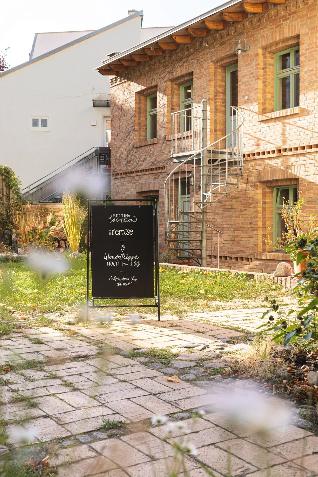 Ein Aufsteller im Garten vor einem roten Backsteingebäude mit grünen Fenstern und einer Wendeltreppe. Der Aufsteller hat handgeschriebene Informationen zu einem Treffen auf Deutsch.