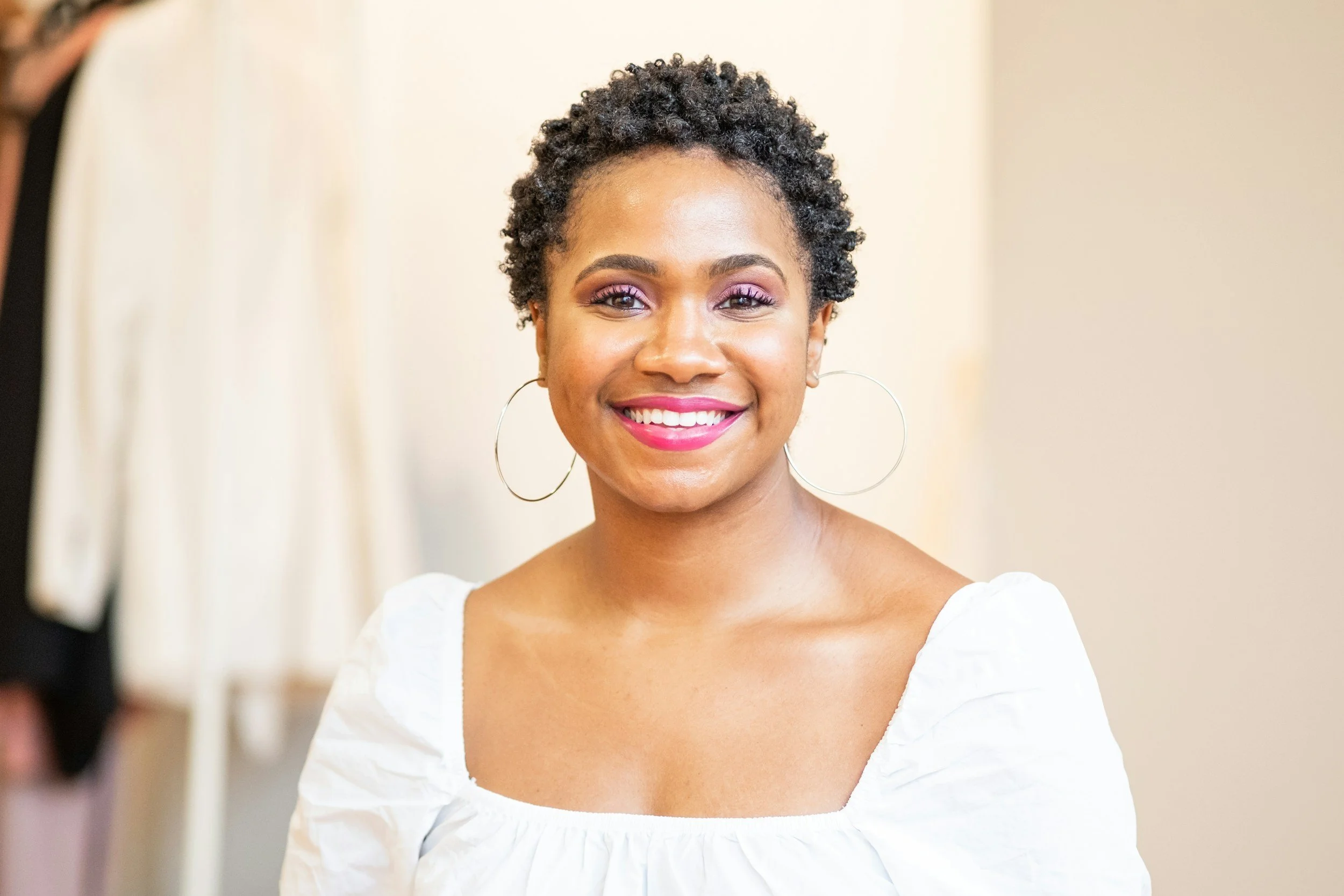 A woman with styled short curly hair and hoop earrings smiling while looking into a mirror, wearing a white top.
