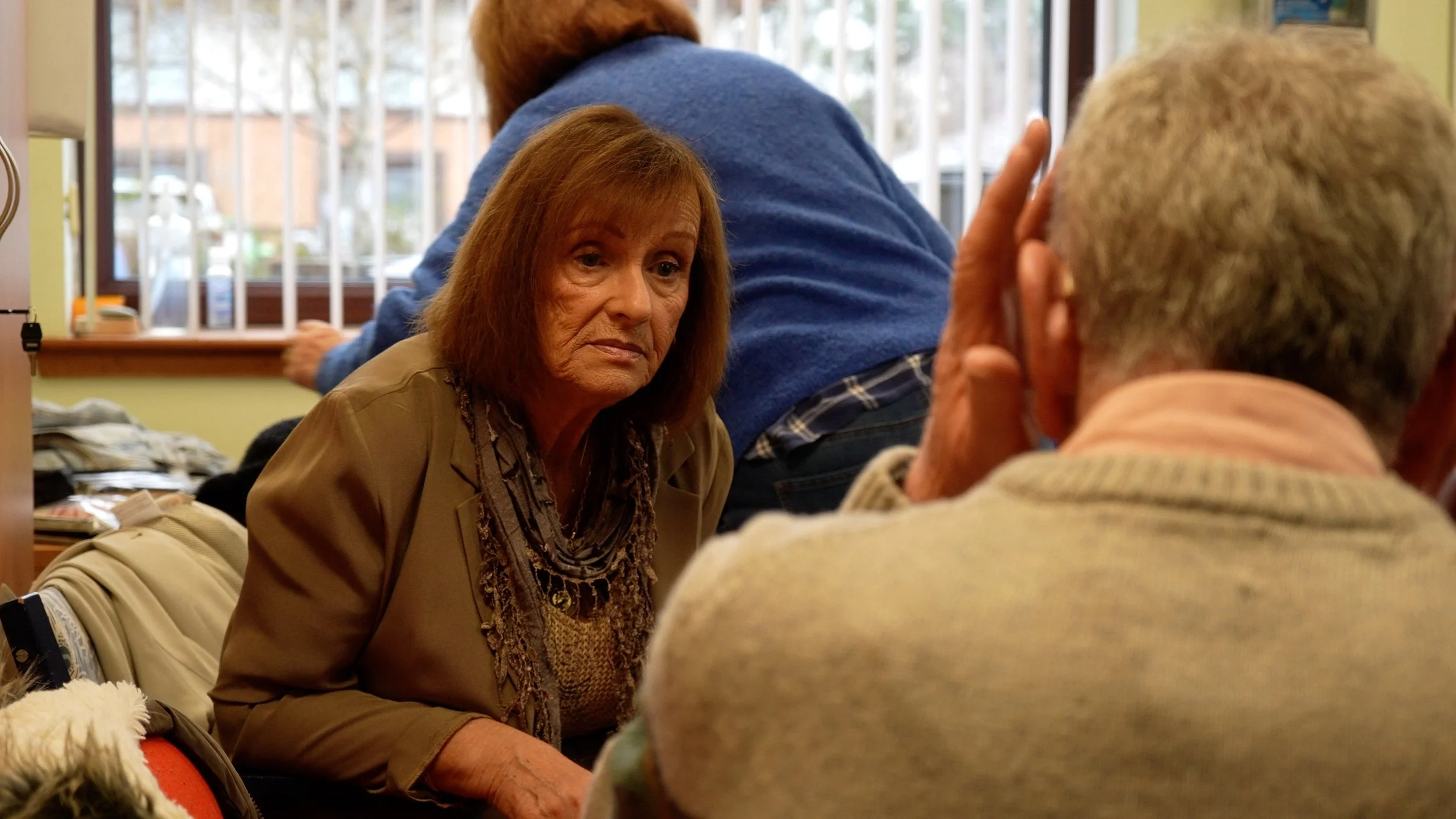 An elderly woman and man engaged in conversation, sitting indoors with a window in the background. The woman has reddish-brown hair and is wearing a jacket and scarf, while the man has gray hair and is wearing glasses and a light-colored sweater.
