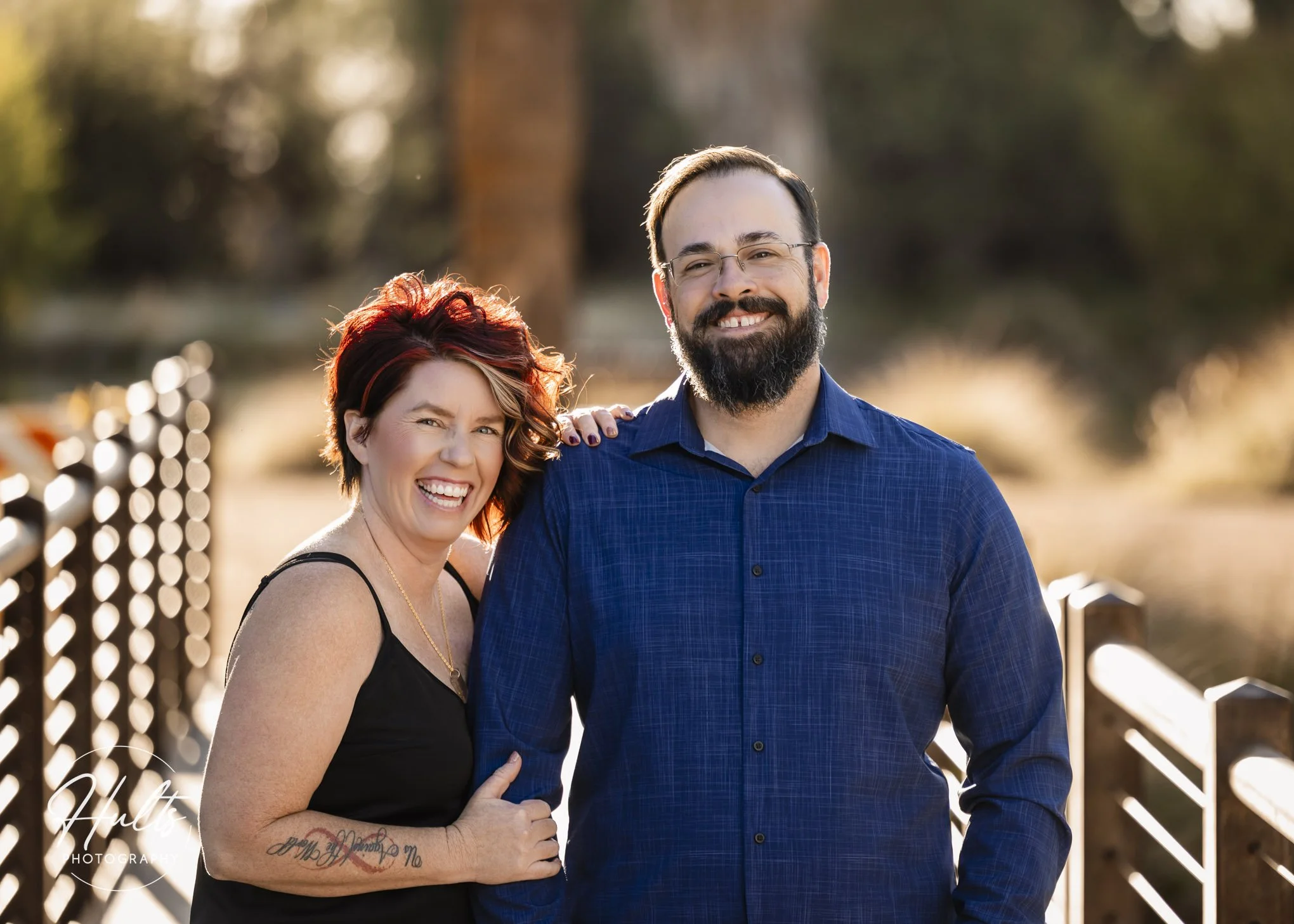 A smiling woman winking and a smiling man standing outdoors by a fence with trees in the background.