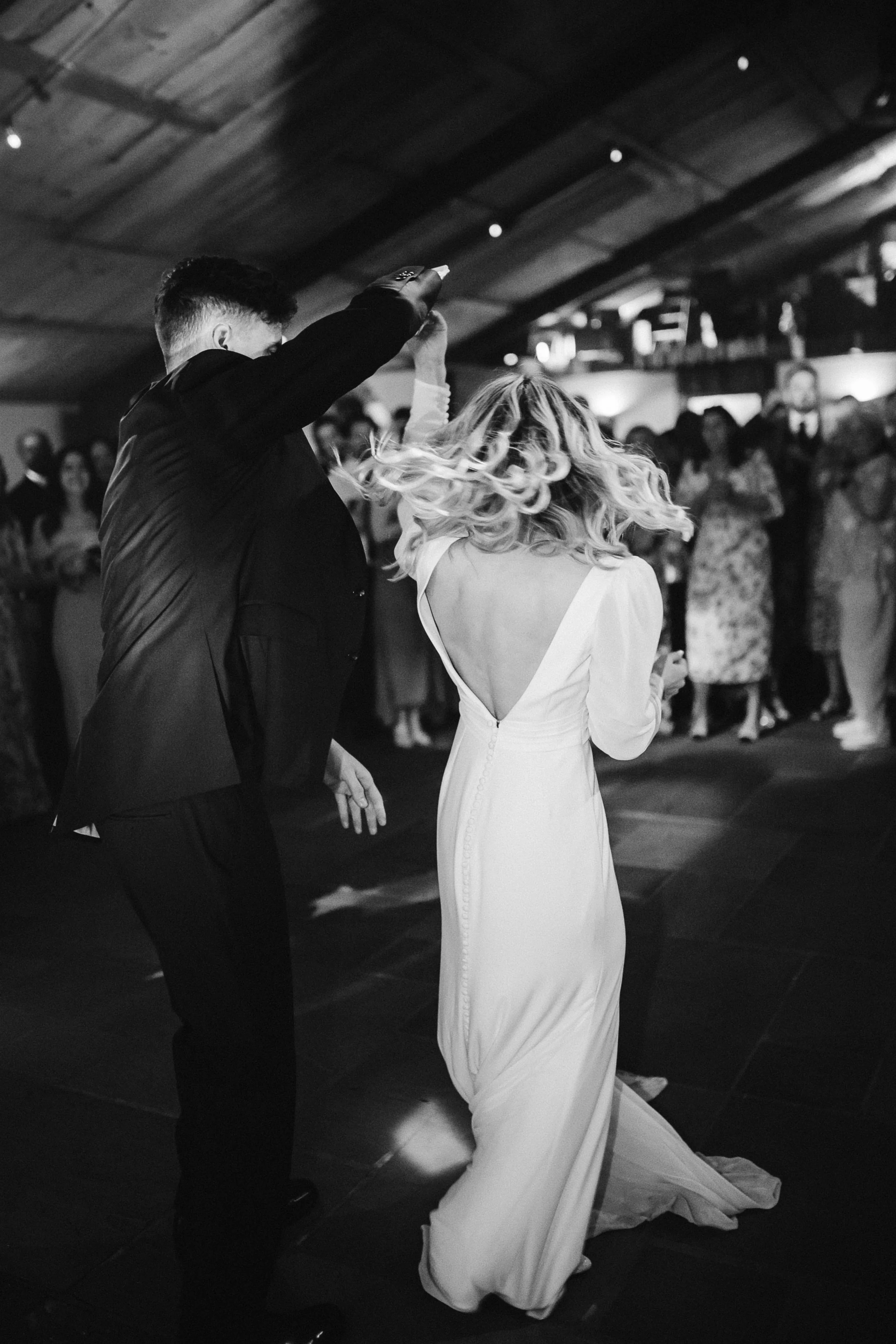 A couple dancing at a wedding reception, with the man raising his hand and the woman twirling in a white gown, while a group of guests watches in the background in a dimly lit indoor setting.