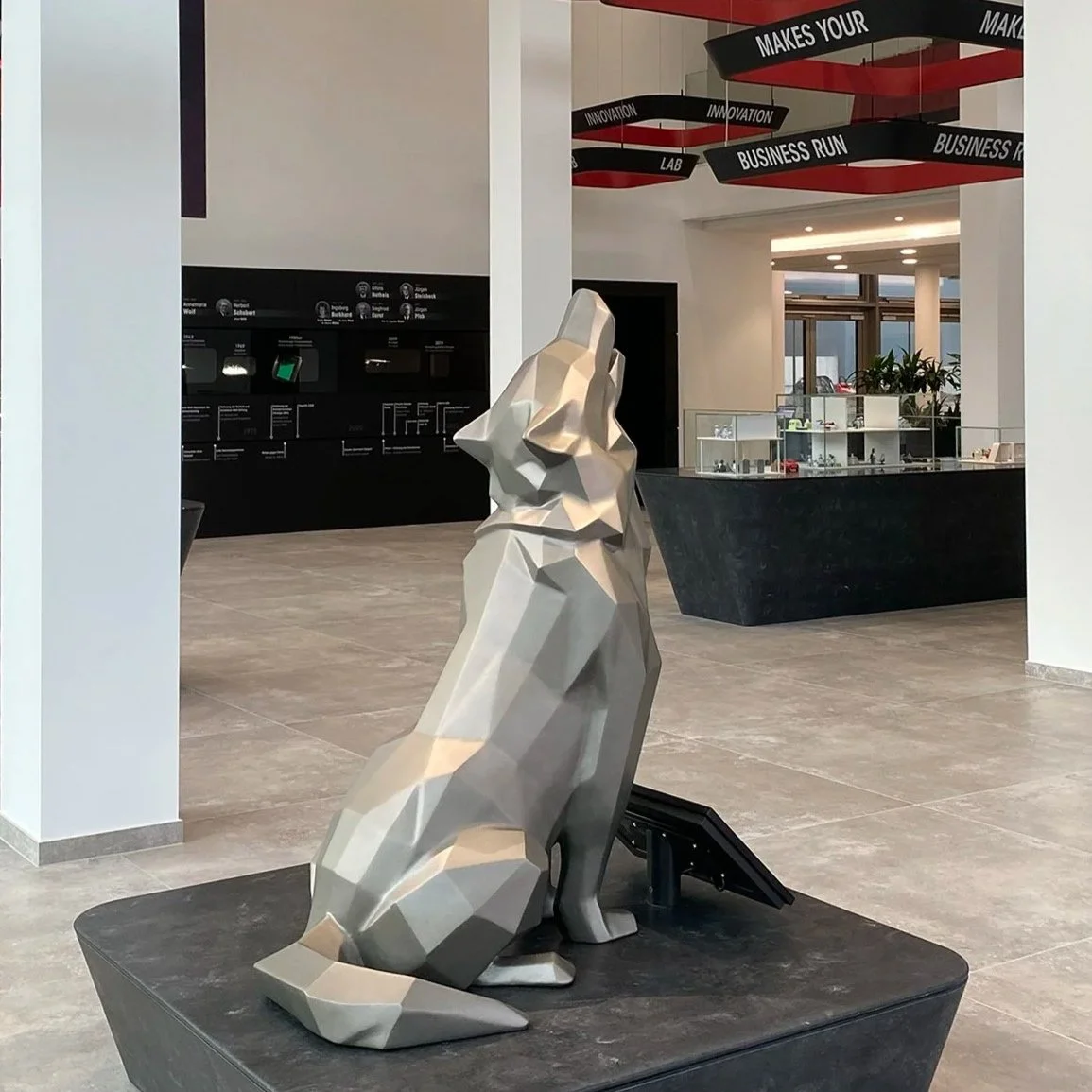 Eine moderne, geometrische Skulptur eines sitzenden Tieres, wahrscheinlich eines Hundes, aus silberfarbenem Metall auf einem schwarzen Sockel in einem modernen Innenraum.