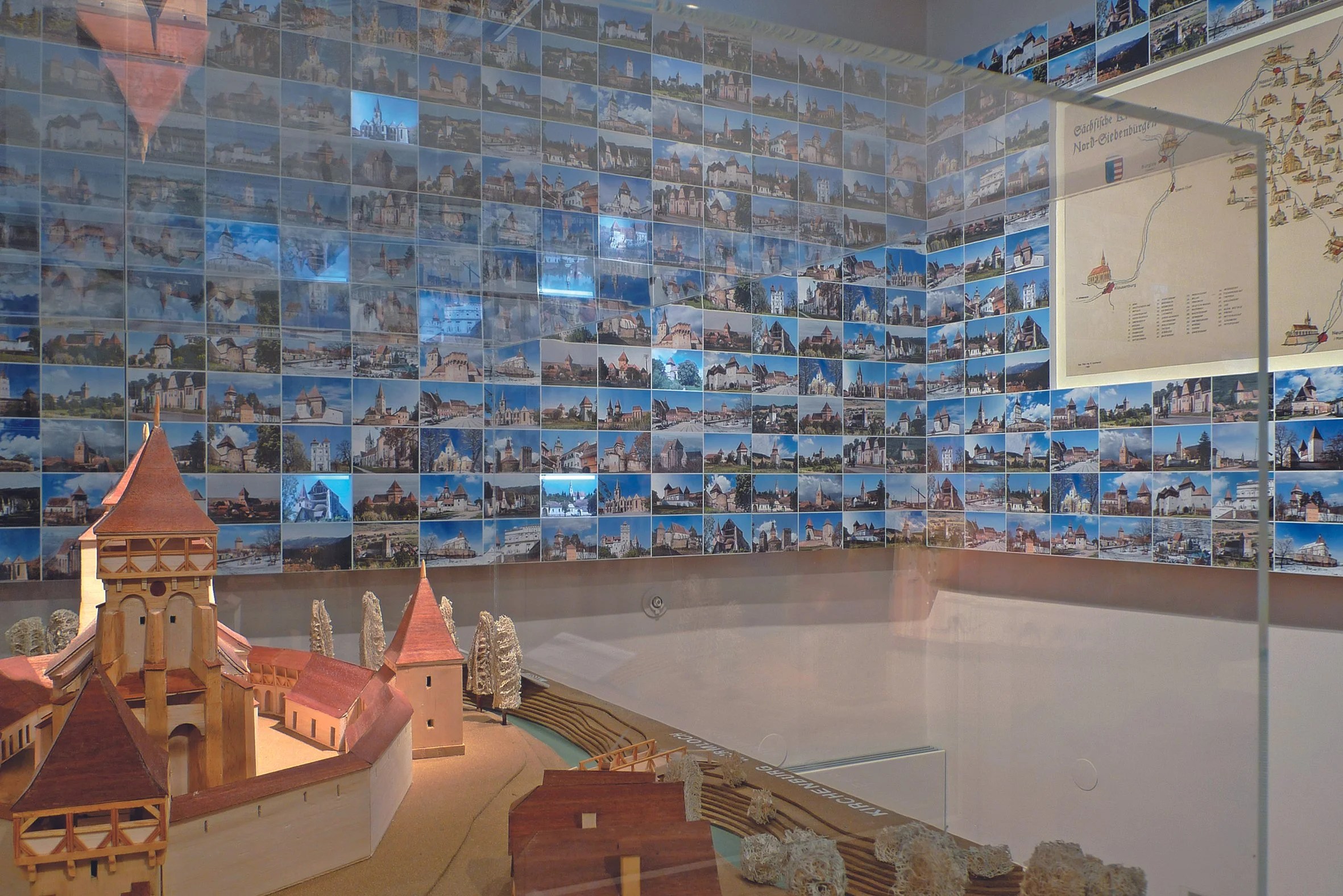 Modell eines historischen Ortes mit mehreren kleinen Gebäuden und einem Wasserkanal, umgeben von einer Wand mit zahlreichen Fotografien und einer Karte an der Wand.