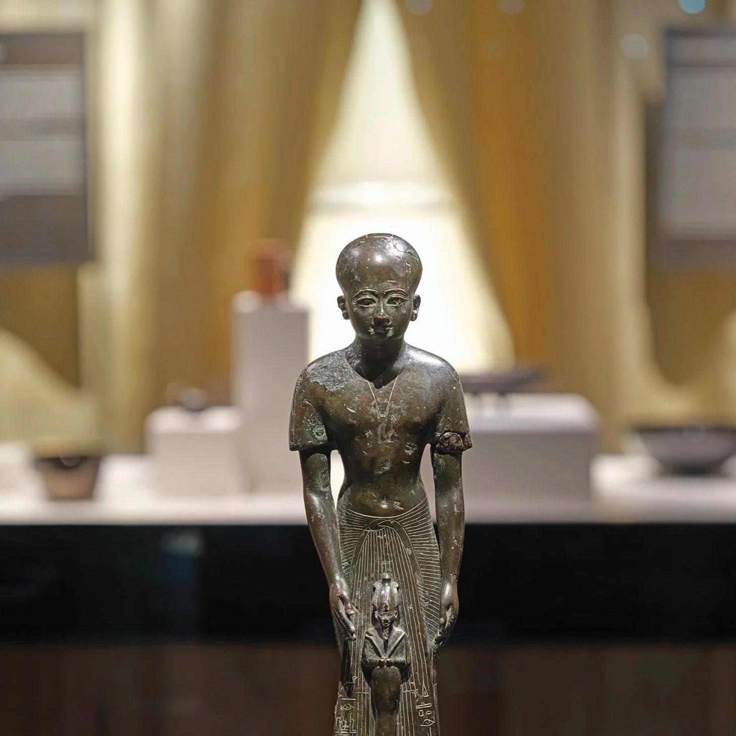 Bronzefigur eines ägyptischen Jungen mit einem Totenkopf, der eine Figur hält. Im Hintergrund ein goldener Vorhang.