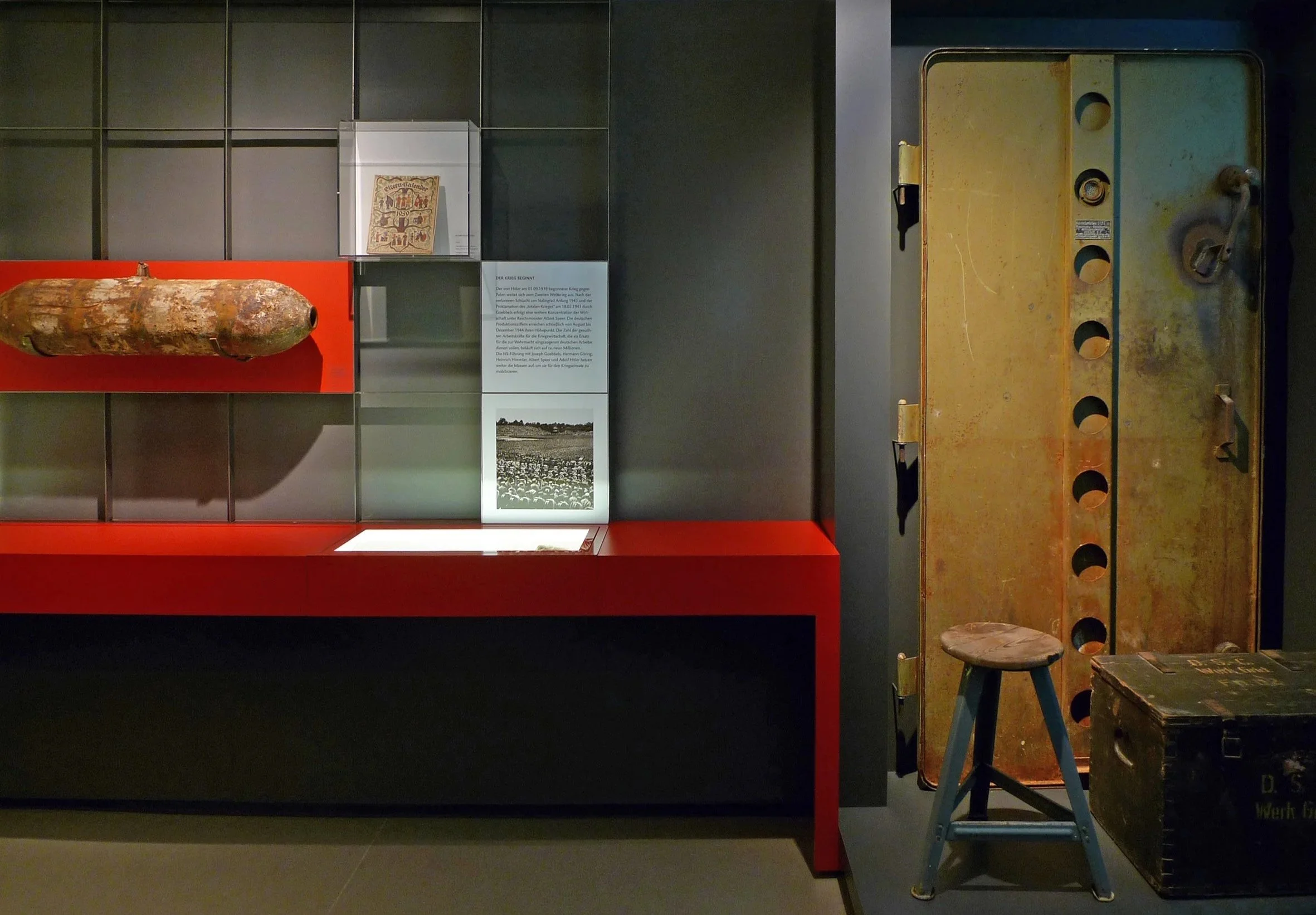 Das Bild zeigt eine Wand mit mehreren Museumsobjekten, darunter eine Bombe, eine Informationsplakette, eine alte Kiste, eine kleine Bank und einen großen, rostigen Metallvorrichtung.