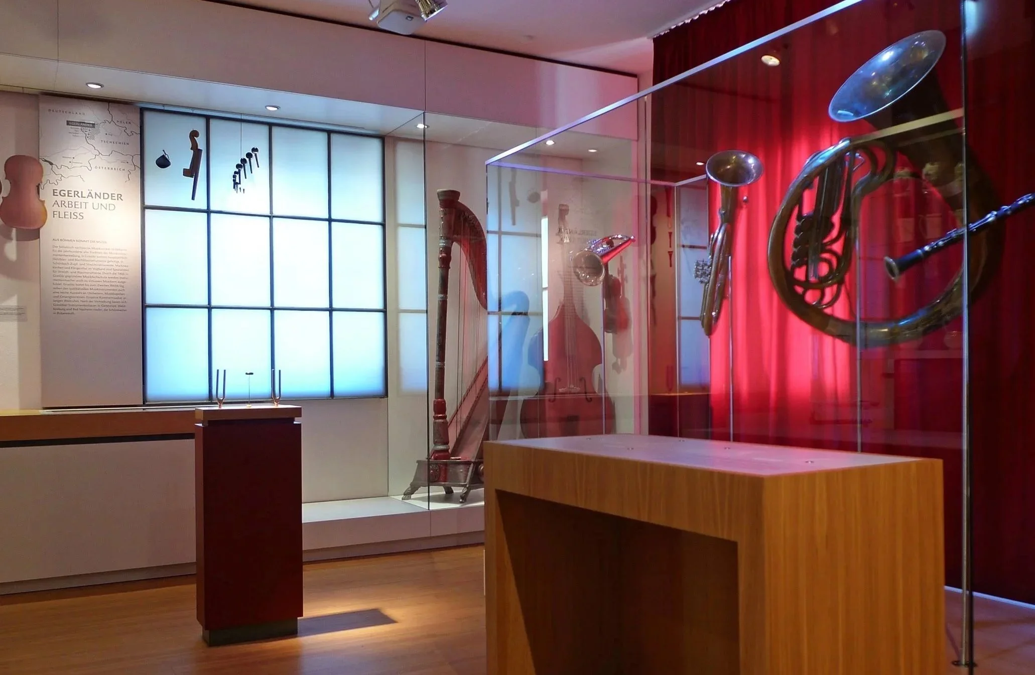 A museum exhibition with musical instruments, including a brass horn, a string instrument, and a wind instrument in a glass display case, with a red curtain in the background.