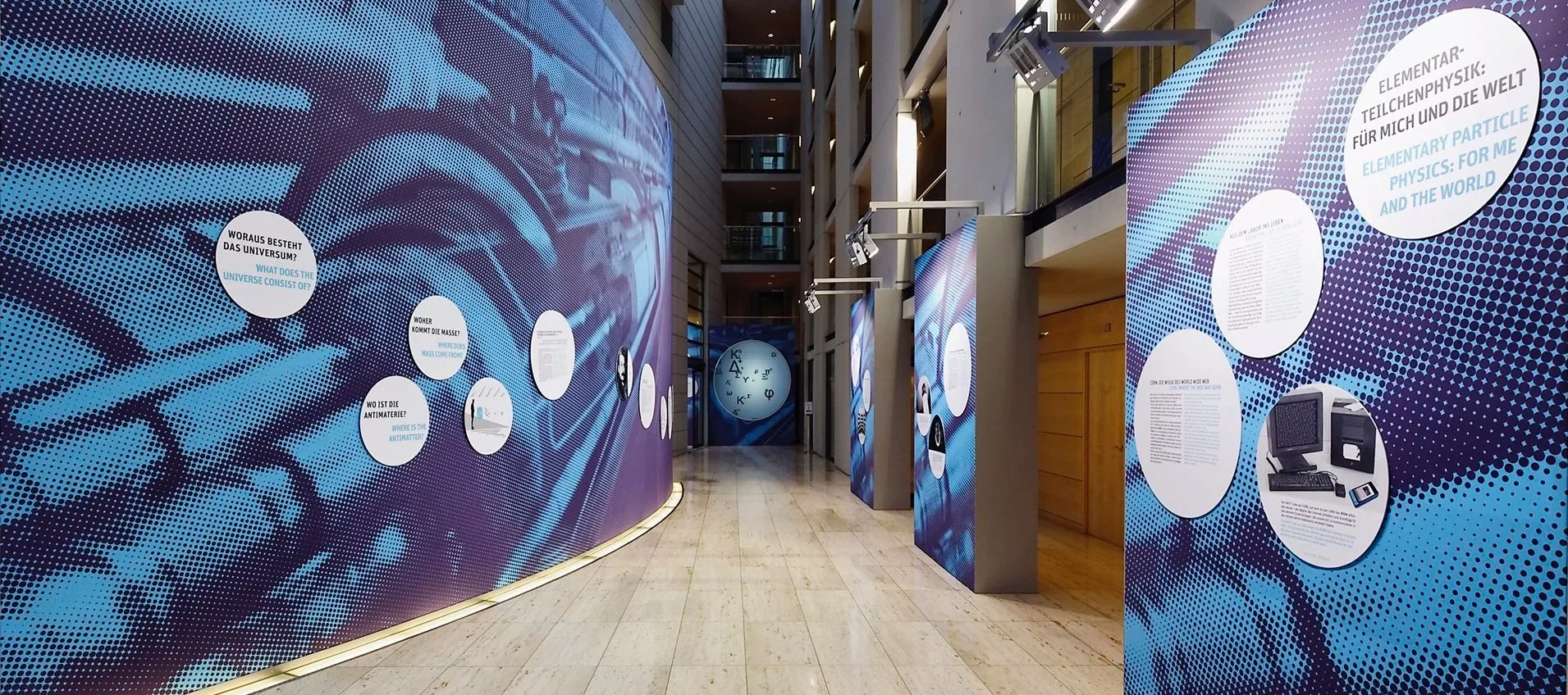 Ausstellungshalle mit blauen digital gestalteten Wänden, auf denen verschiedene Texte in Deutsch und Englisch zu Themen wie Universum, Masse, Antimaterie und Physik angezeigt werden. Der Raum ist modern gestaltet mit Holztüren und metallischen Stände