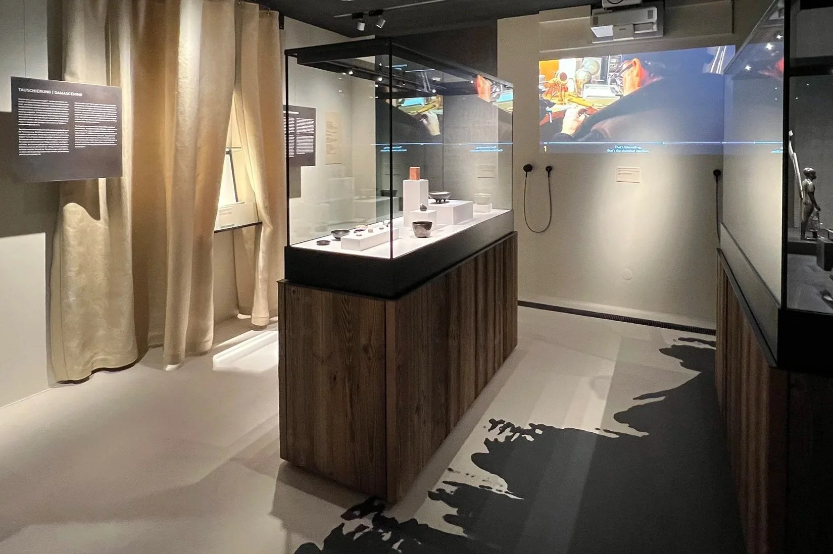 Virtuelles Museum mit einer Glasvitrine, die antike Keramik in einem schlichten Raum zeigt, im Hintergrund ist eine TV-Demo, die den Herstellungsprozess zeigt.