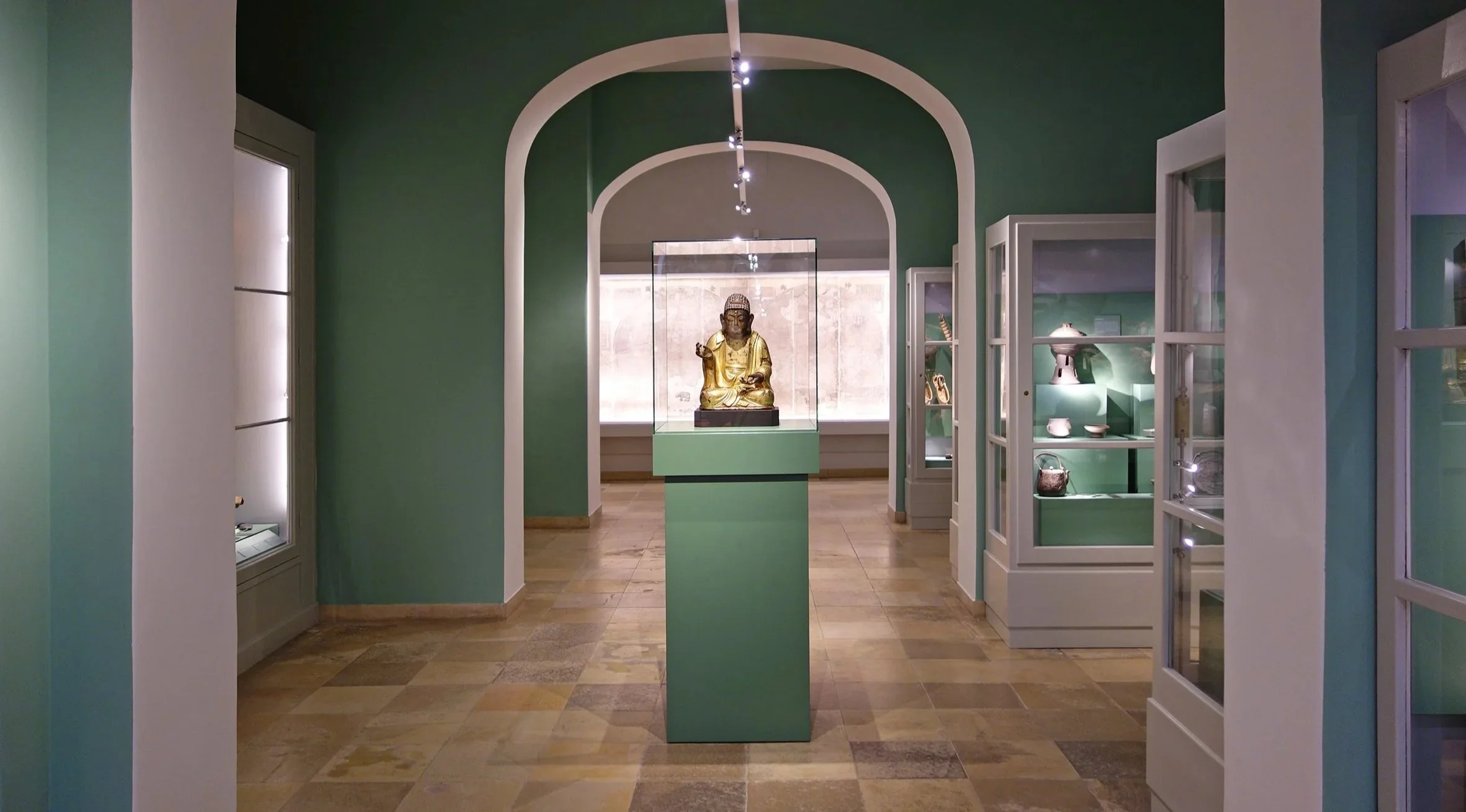 Blick in ein Museum mit einer goldenen Statue in einer Glasvitrine in der Mitte. Der Raum hat grüne Wände und mehrere Glasvitrinen mit Artefakten. Der Boden ist aus Holzparkett.