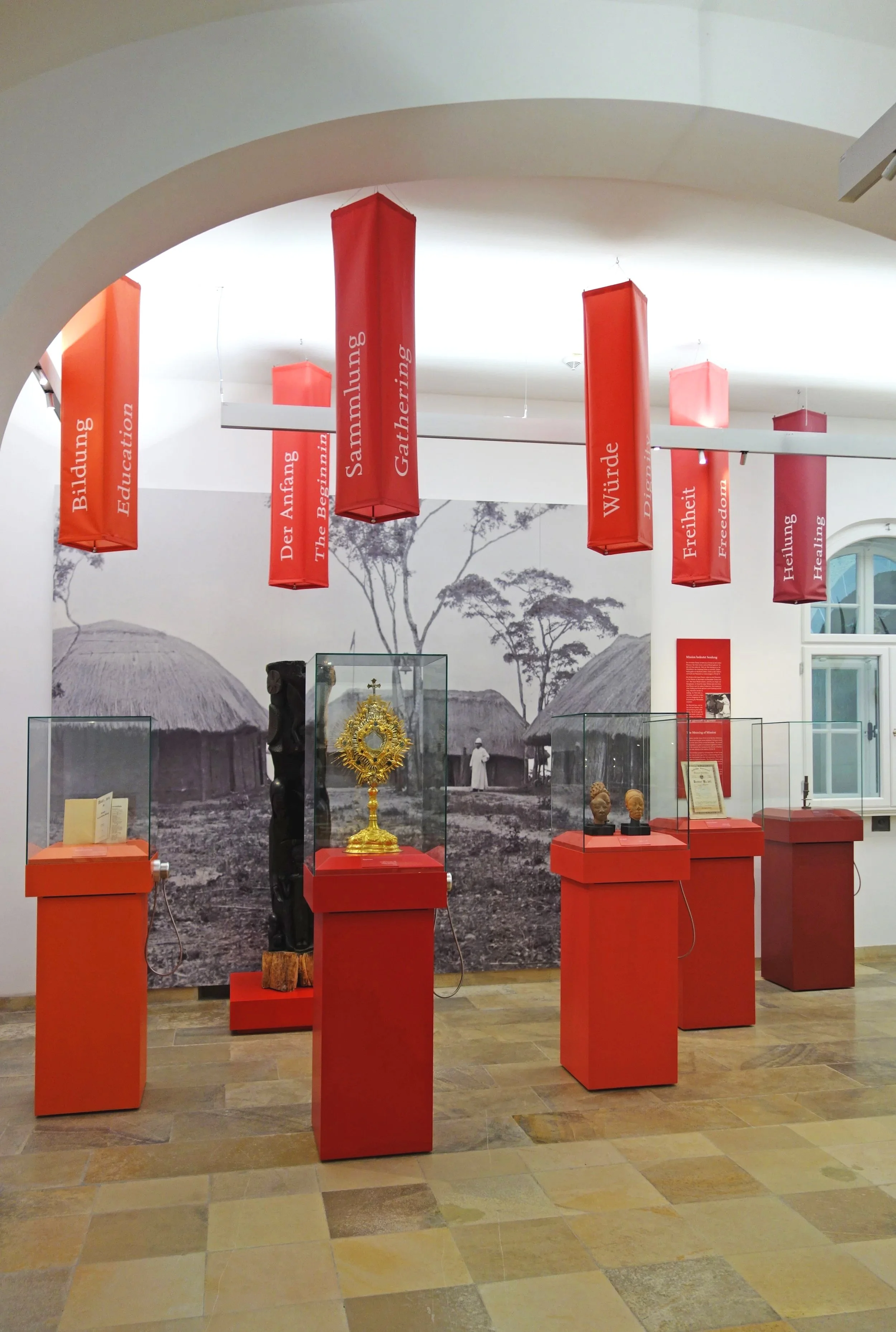 Ausstellung im Museum mit roten Podesten und Glassäulen, darin kunstvolle Objekte wie eine goldene Monstranz und Antikenfiguren. Die Wand im Hintergrund zeigt eine schwarze und weiße Fotografie eines Dorfes mit Rundhütten und Menschen, sowie rote Sch