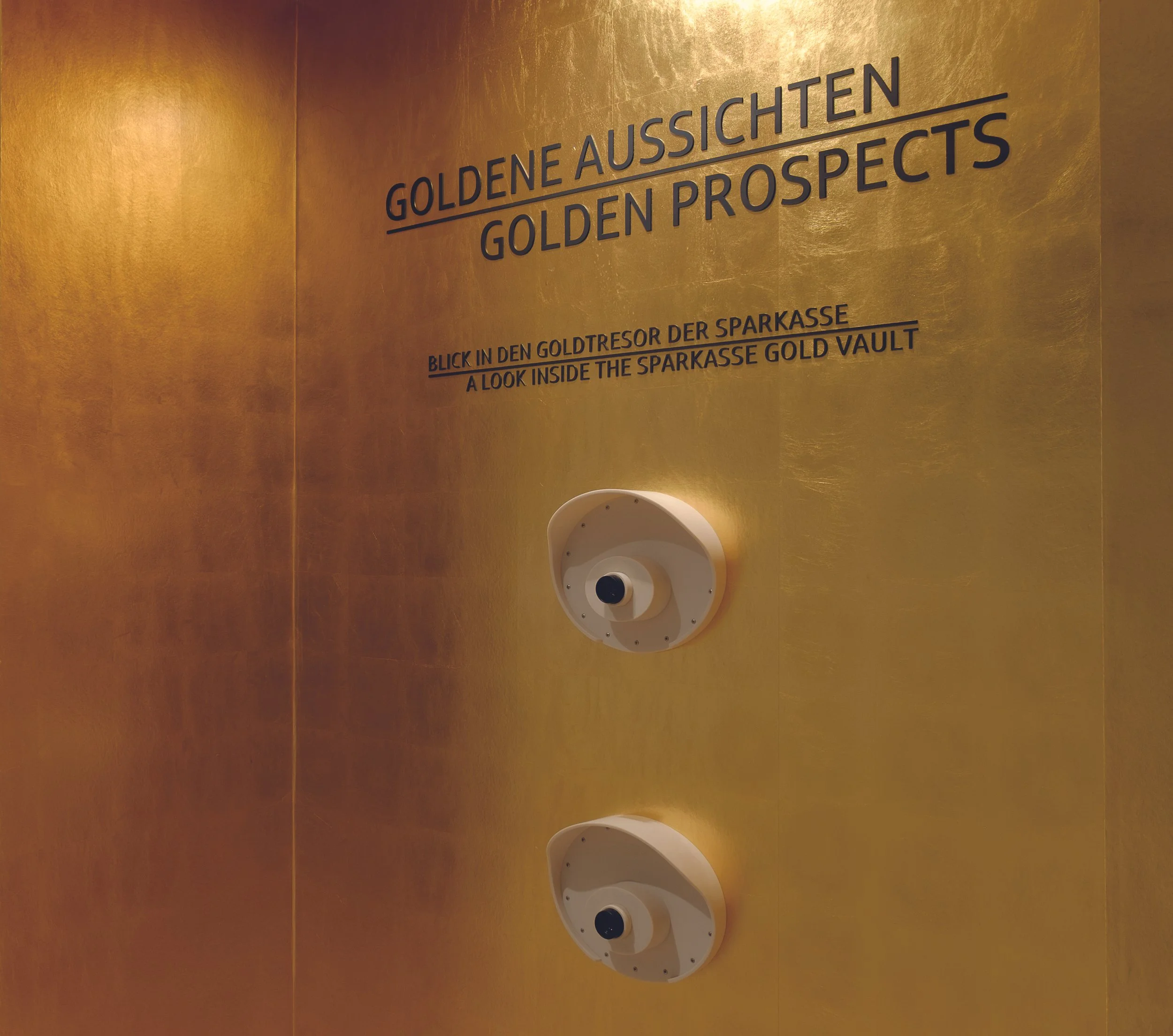 Blick in den Goldtresor der Sparkasse mit goldener Wand und Ausdruck 'GOLDENE AUSSICHTEN' sowie 'GOLDEN PROSPECTS' in großen Buchstaben, und zwei runde Sicherheitsfenster.