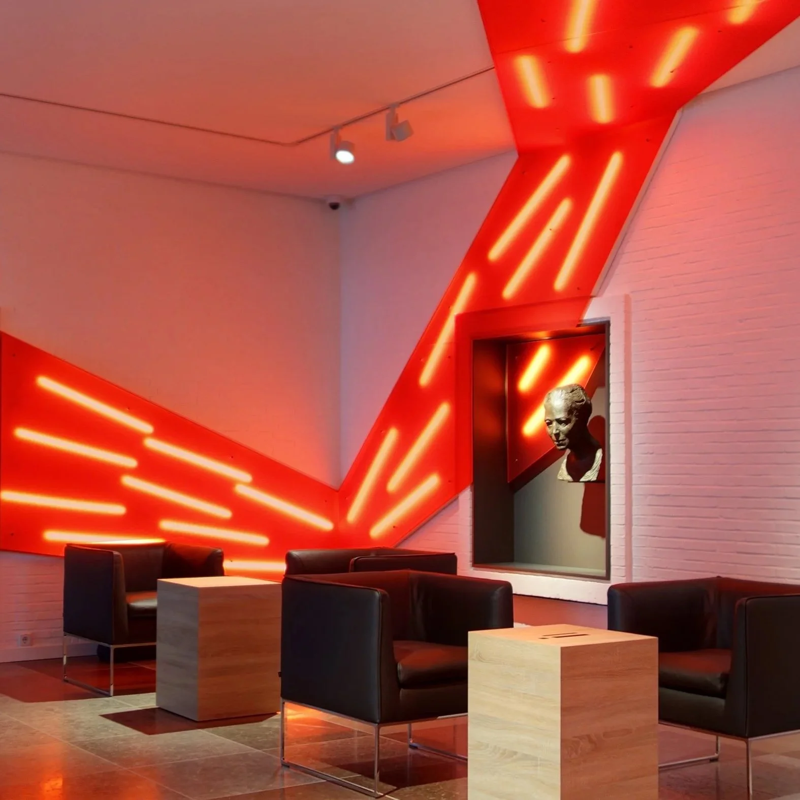 Modernes Wartezimmer mit schwarzen Ledersesseln, weißen Tischen und einer Wand mit roten Neonlichtern. Ein schwarz-weißer Porträtkopf ist in einer Nische an der Wand zu sehen.