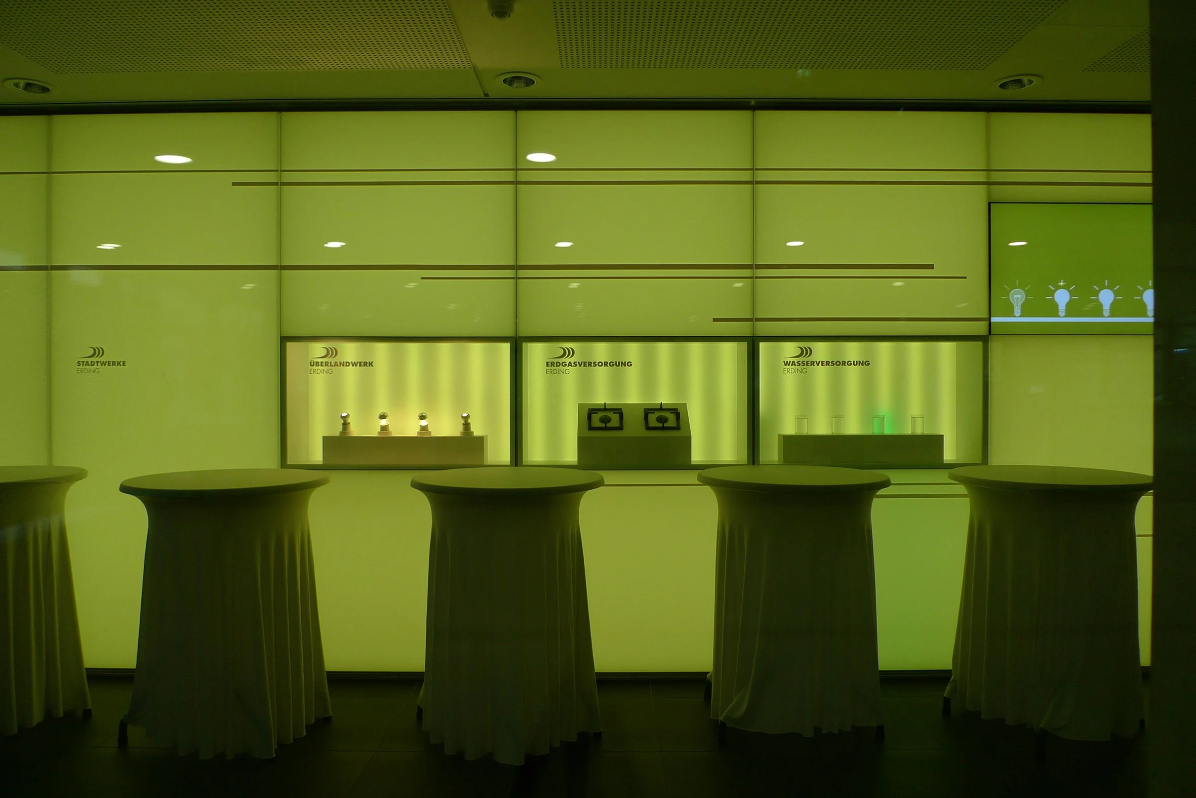Ein hellgrüner Hintergrund mit vier weißen Stehtischen im Vordergrund. Dahinter befinden sich drei Abschnitte mit verschiedenen Wasser- und Gasanschlüssen, die mit deutschen Beschriftungen wie "Stadtwerke Erdingen", "Überlandwerk Erding" und "Erdgasv