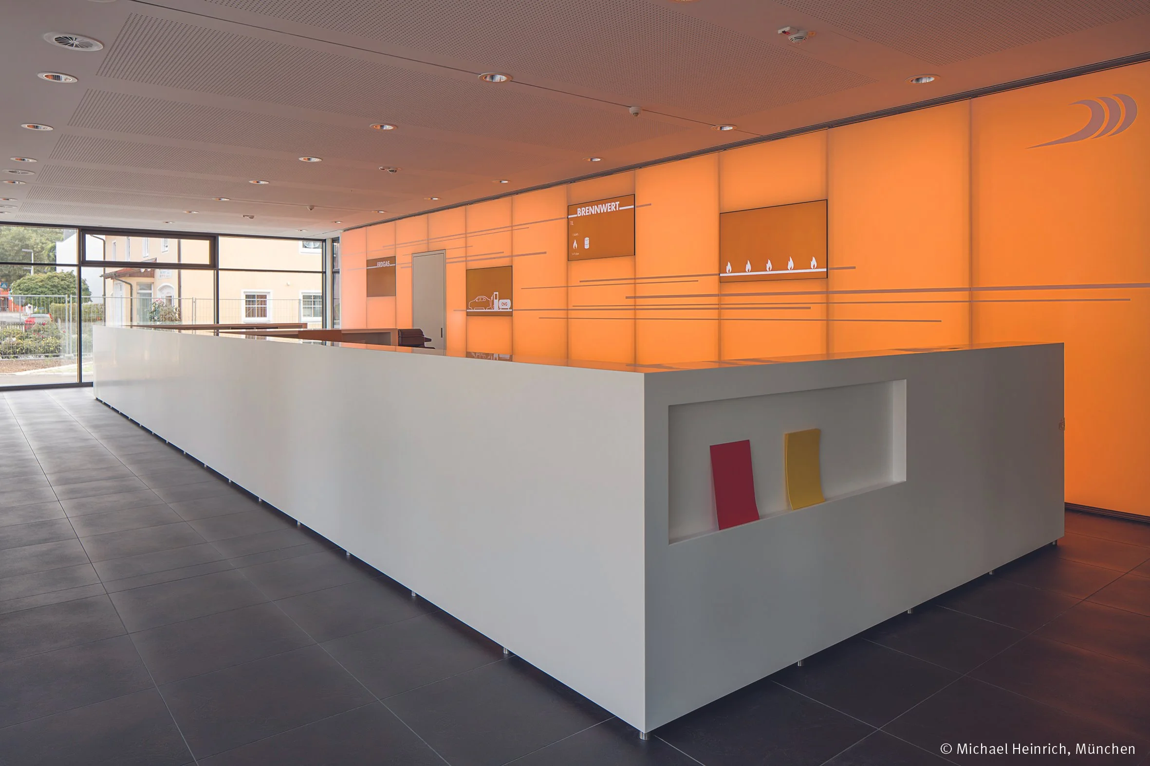 Moderne Empfangstheke in einem hell beleuchteten Raum mit großen Fenstern, orangefarbener Wanddekoration, und einer Ablagefläche. Es gibt einige Schriften und Symbole an der Wand sowie eine eingebaute Regaleinheit mit bunten Zeitschriften.